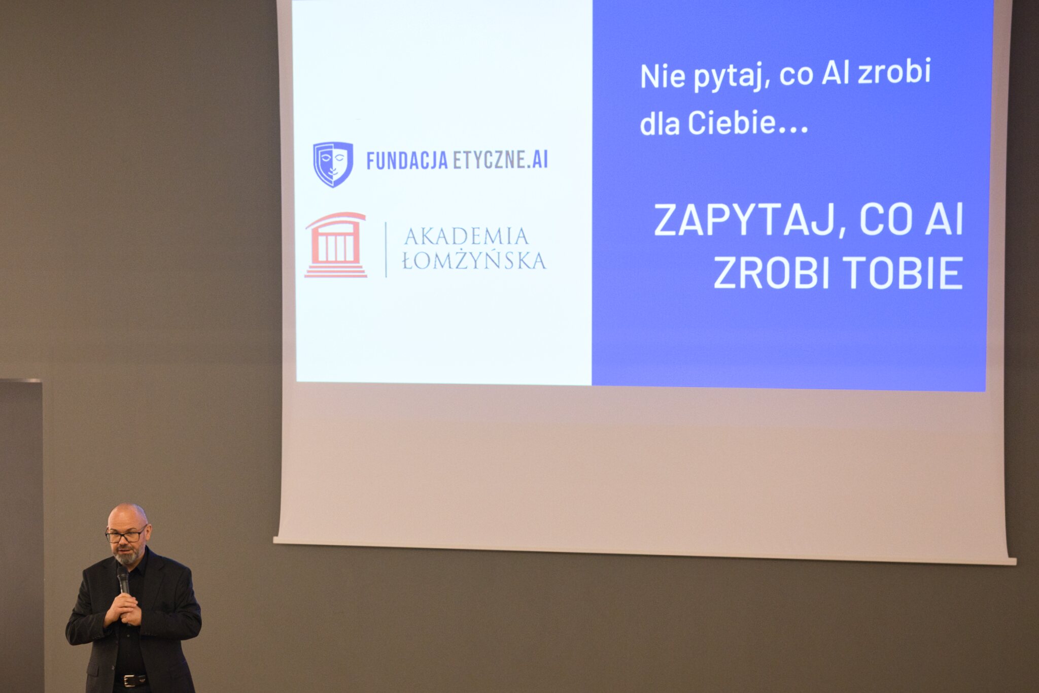Osoba prowadząca wykład mówi przez mikrofon, za nią wyświetlana jest prezentacja, na której jest napisane "Nie pytaj co AI zrobi dla Ciebie...zapytaj co zrobi Tobie"