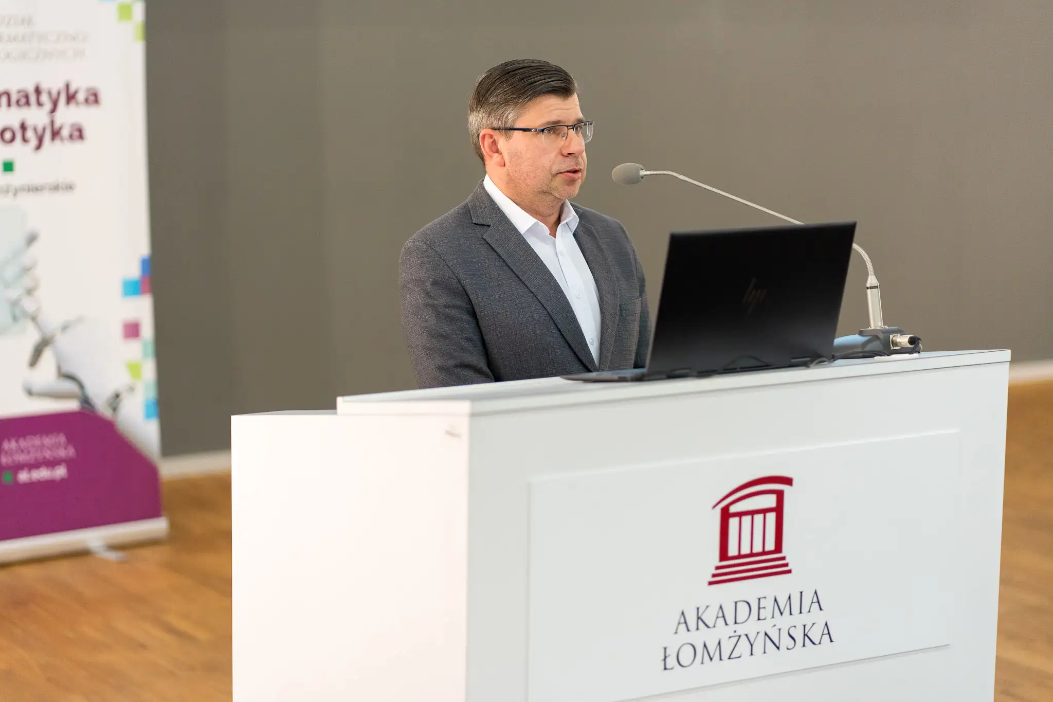 Osoba stoi za białą mównicą z logo Akademii Łomżyńskiej i wygłasza prezentację, korzystając z laptopa ustawionego na pulpicie. Obok mównicy znajduje się mikrofon, a w tle widoczny jest fragment roll‑upu z elementami graficznymi.