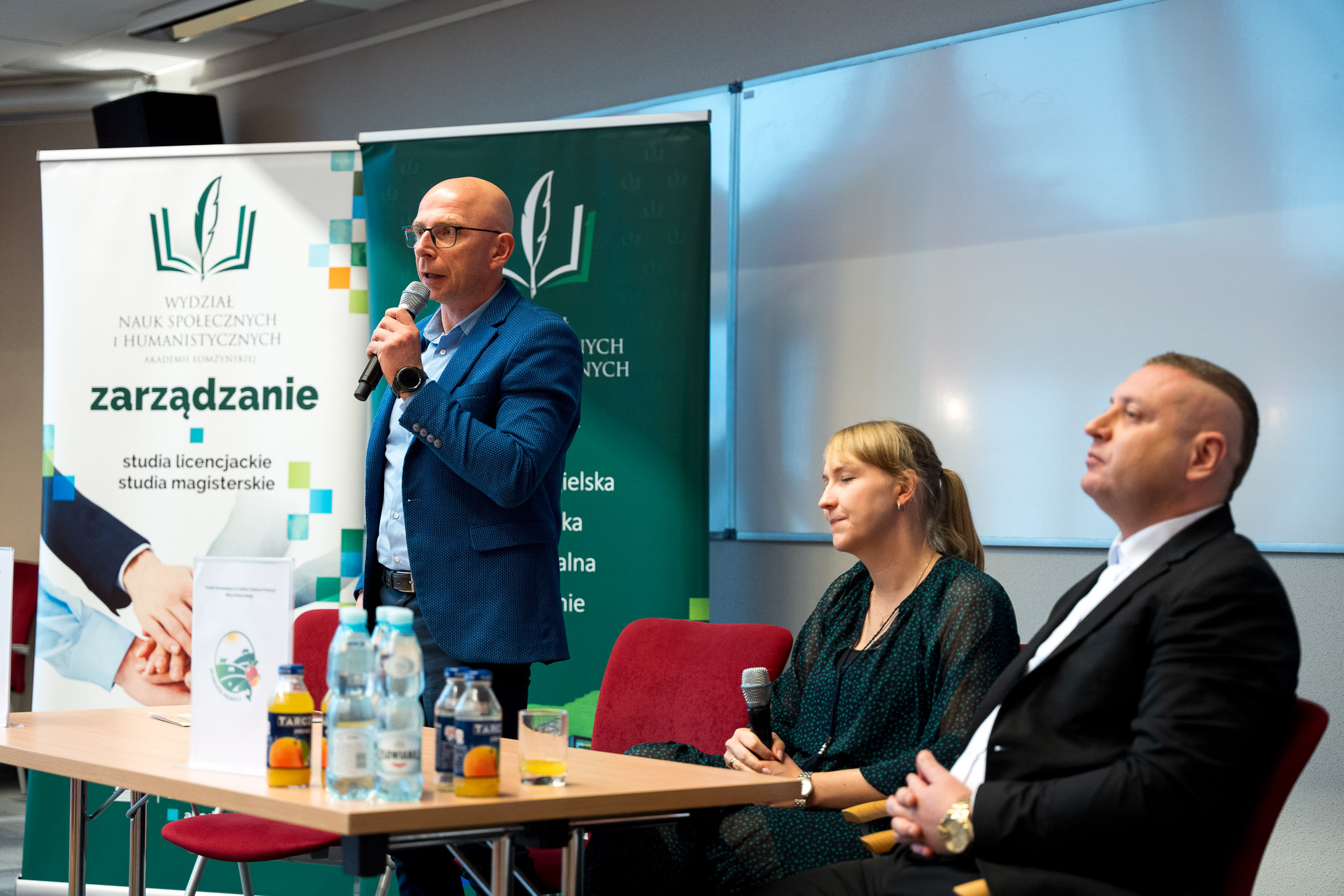 Doktor Adam Miara przemawia w auli 315. Dwórch panelistów siedzi obok.
