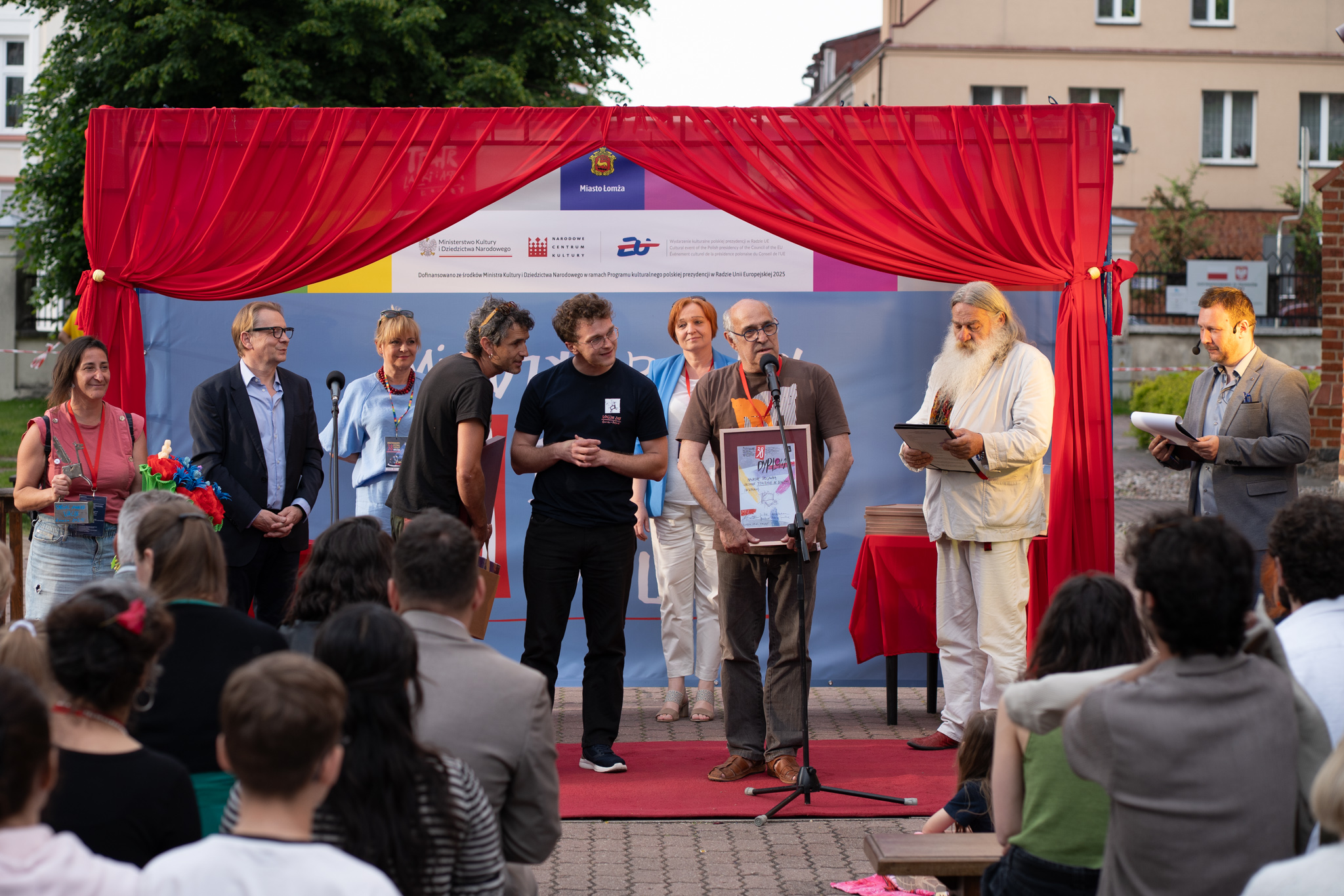 Kadr z wręczania nagród. Widoczne 7 osób. Na pierwszym planie 3 mężczyzn: jeden trzyma nagrodę i mówi do publiczności, drugi mężczyzna tłumaczy na język angielski wypowiedź pierwszego, a trzeci męzczyzna, będący studentem tłumaczy na język polski