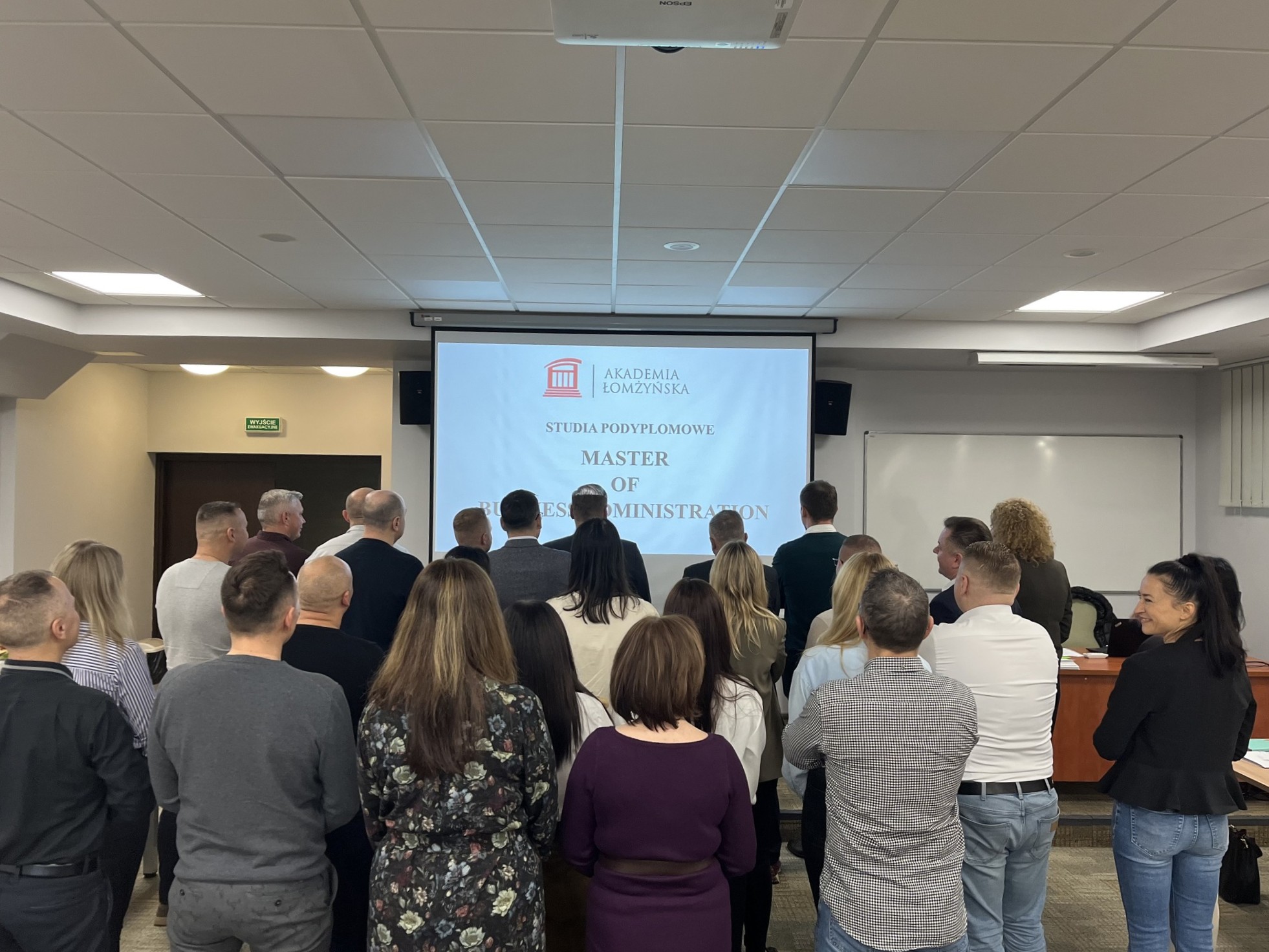 Grupa ludzi stoi w sali ćwiczeniowej. Na drugim planie widoczny zlajd z napisem: MBA Master of business administration i grafiką dekoracyjną