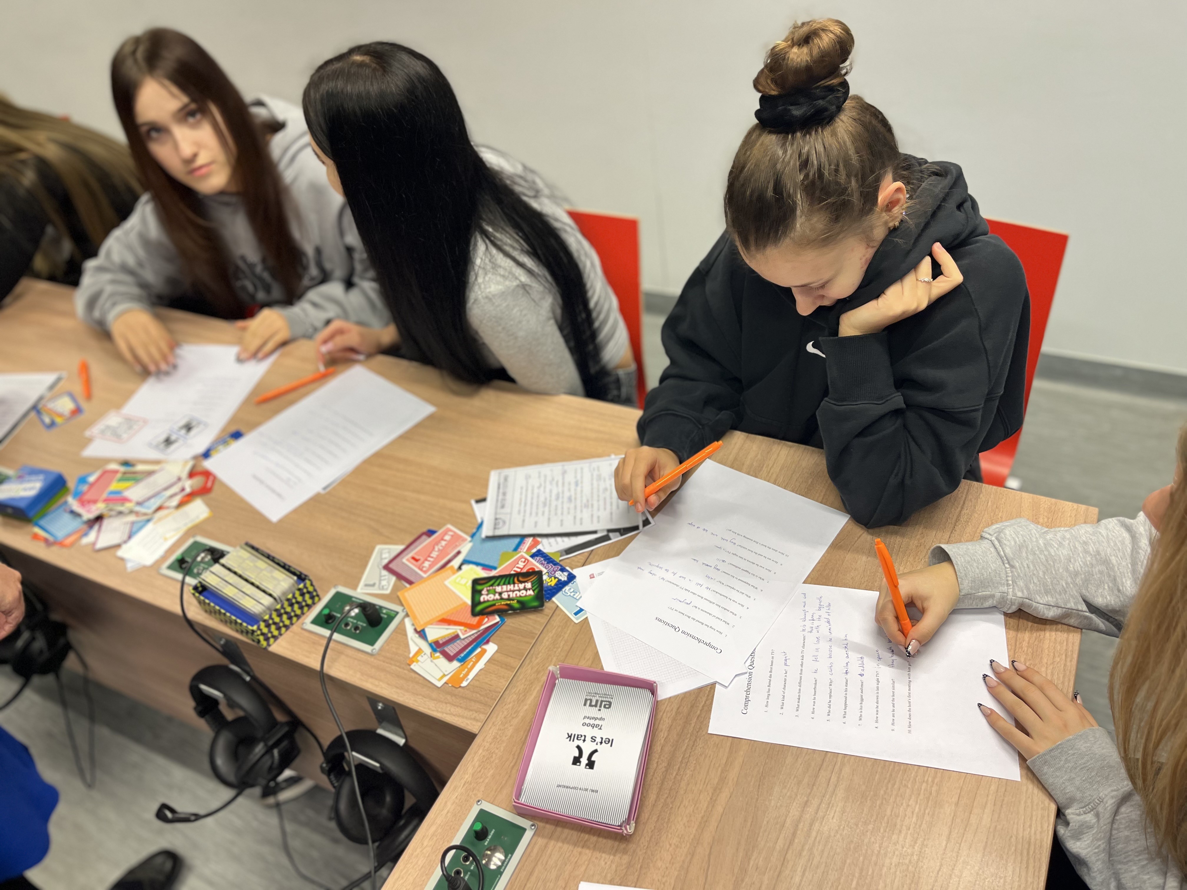 cztery młode dziewczyny siedzą przy ławkach i rozwiązują test językowy. Obok na blacie leżą kolorowe karty do gry z napisami po angielsku