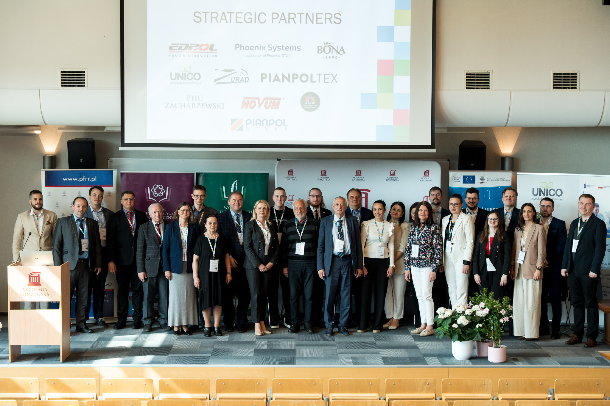 Duża grupa uczestników konferencji pozuje na scenie. W tle slajd z widocznymi logówkami partnerów strategicznych