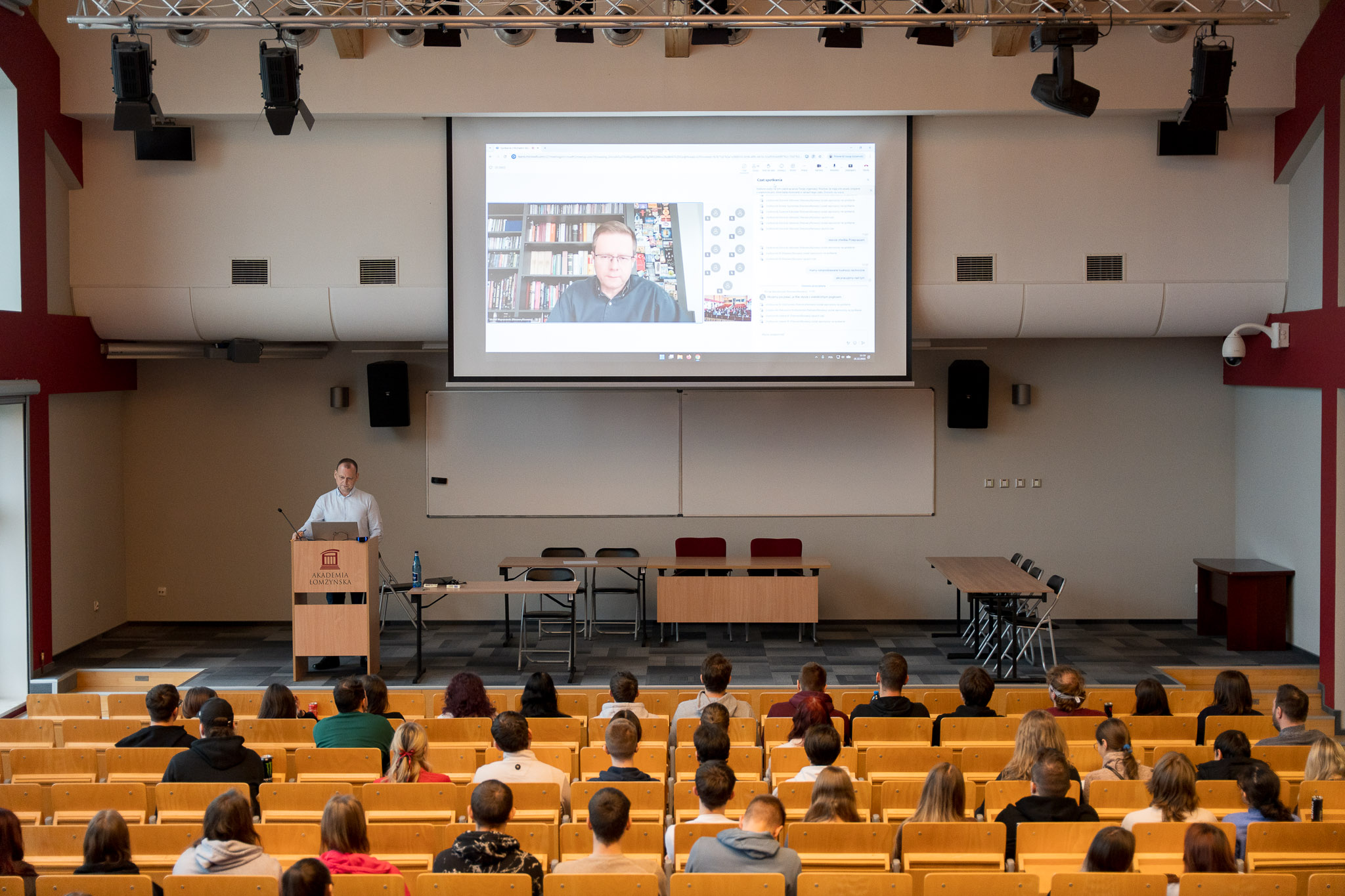 Ujęcie na scenę auli. Na pierwszym planie studenci siedzący na widowni. Na scenie po lewej stronie przy mównicy stoi dr Sitkowski. W centrum sceny na ekranie widoczny zaproszony gość