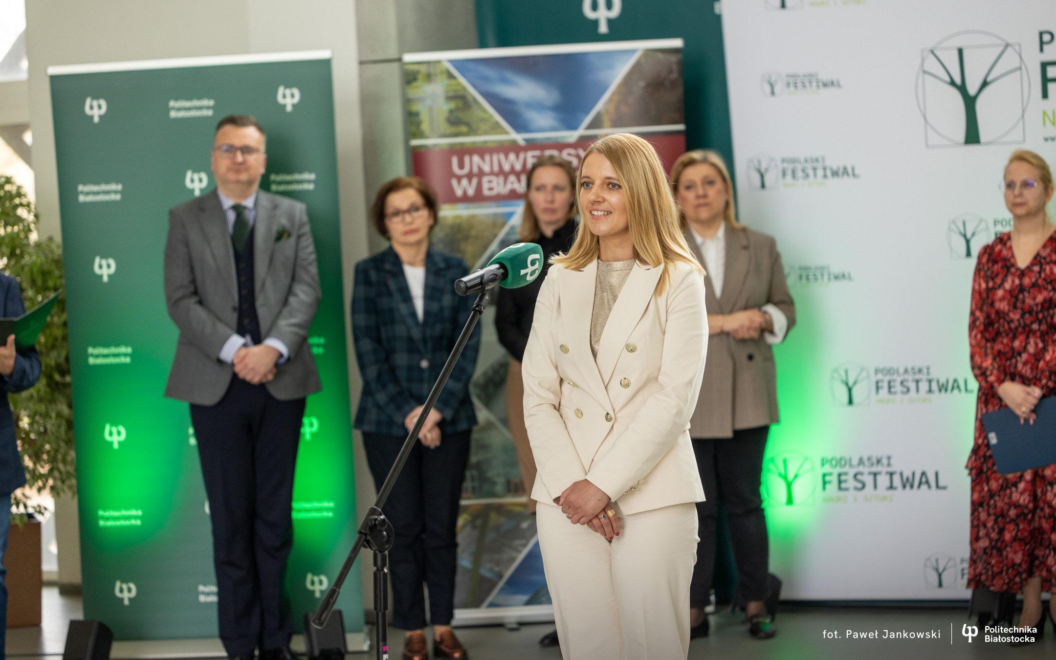 Osoba przemawiająca przy mikrofonie podczas wydarzenia naukowego; w tle stoją uczestnicy oraz banery Politechniki Białostockiej i Podlaskiego Festiwalu Nauki i Sztuki.