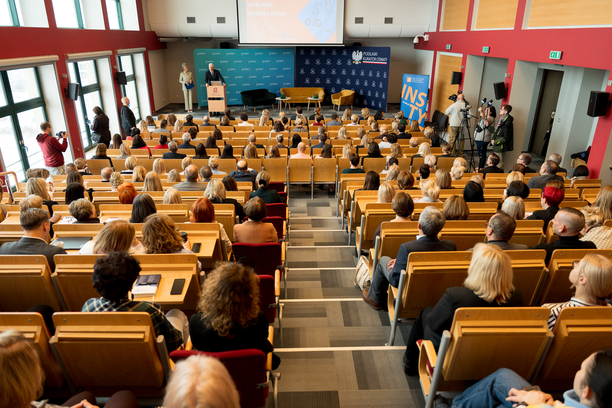 Duża sala wykładowa z licznie zgromadzonymi uczestnikami siedzącymi w rzędach, słuchającymi wystąpienia osoby stojącej przy mównicy na scenie