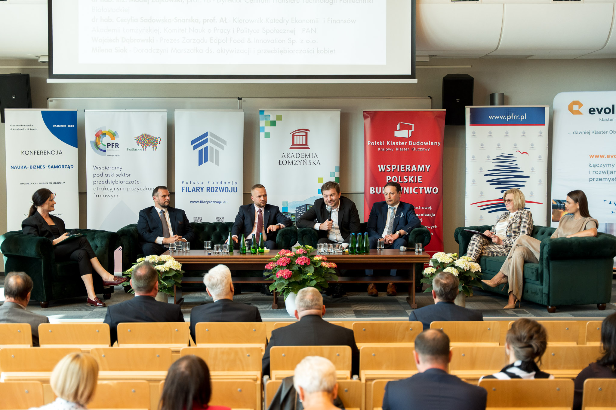 Panel dyskusyjny podczas konferencji w sali wykładowej, z kilkoma prelegentami siedzącymi na scenie i publicznością w pierwszych rzędach.