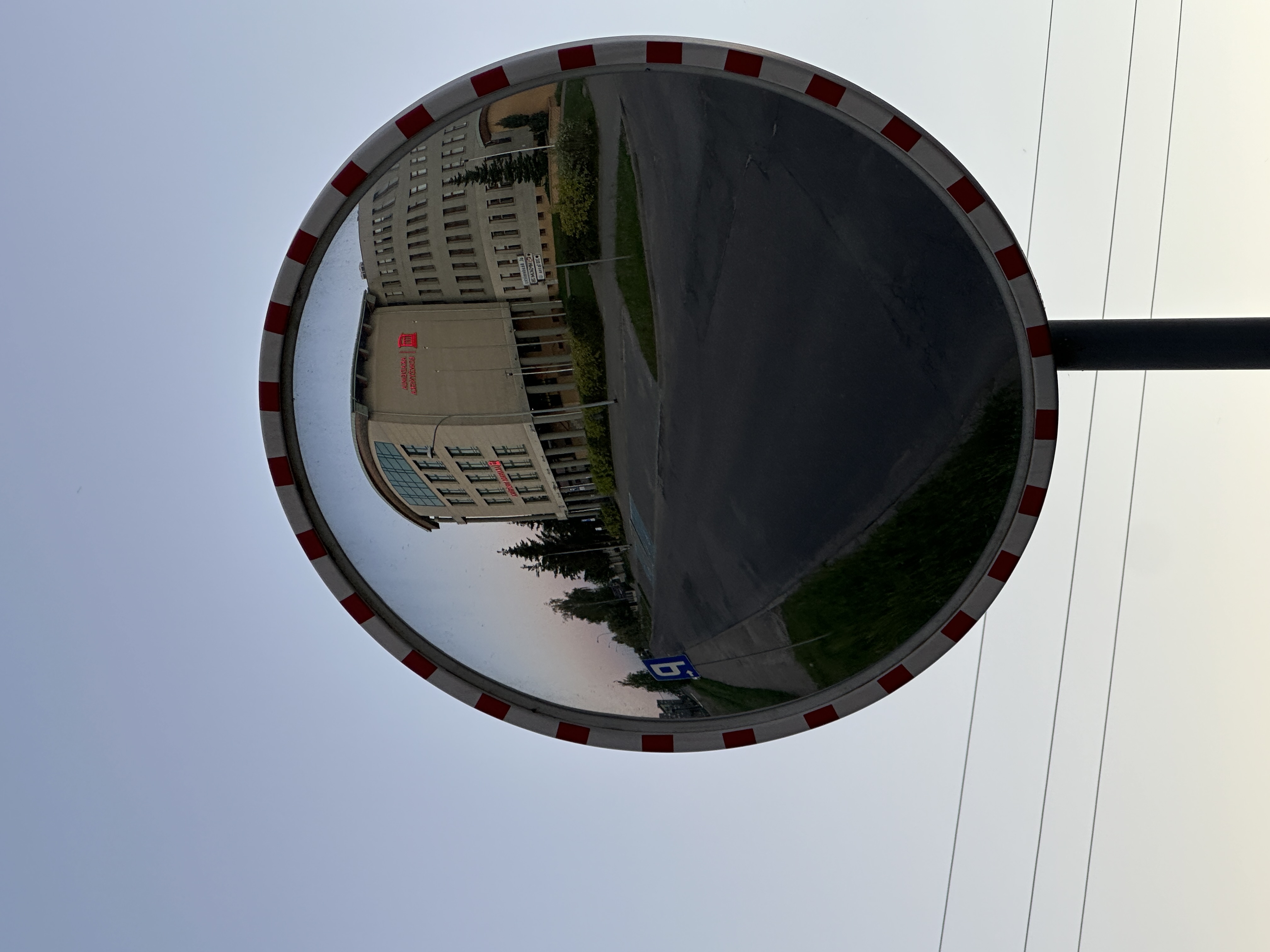 Zdjęcie przedstawia duże, okrągłe lustro drogowe zamontowane na słupie, z czerwono-białą ramką. W odbiciu lustra widoczny jest fragment ulicy oraz nowoczesny, jasny budynek o zaokrąglonym narożniku i wielu oknach. Na elewacji budynku znajduje się czerwony napis oraz logo, jednak szczegóły tekstu są trudne do odczytania.