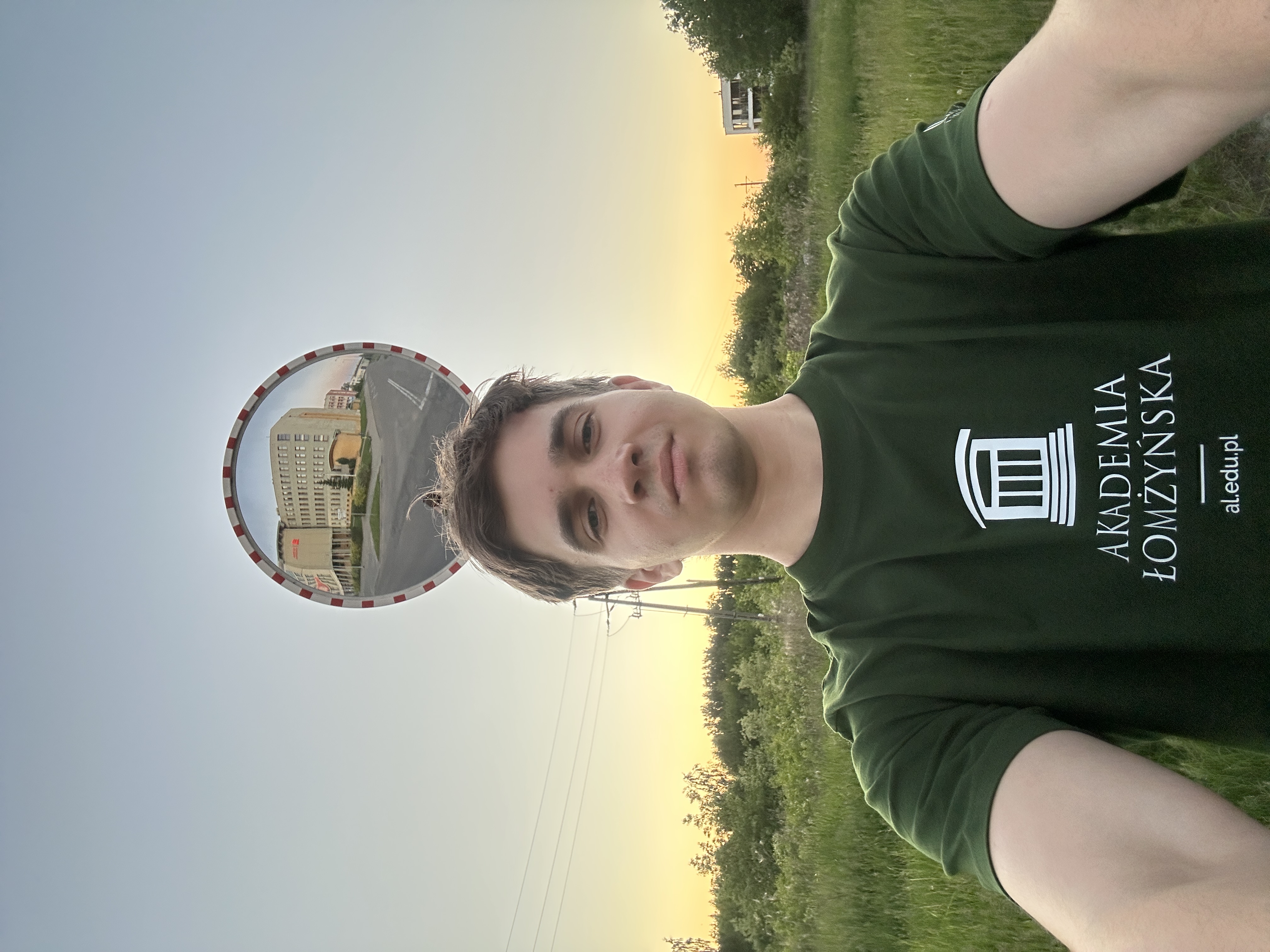 Zdjęcie typu selfie wykonane na zewnątrz, przedstawiające osobę znajdującą się na pierwszym planie, ujętą od ramion w górę. Osoba trzyma aparat lub telefon przed sobą, patrząc w jego stronę. Ubrana jest w ciemnozieloną koszulkę z białym nadrukiem zawierającym napis „Akademia Łomżyńska” oraz logo w formie stylizowanej kolumny.