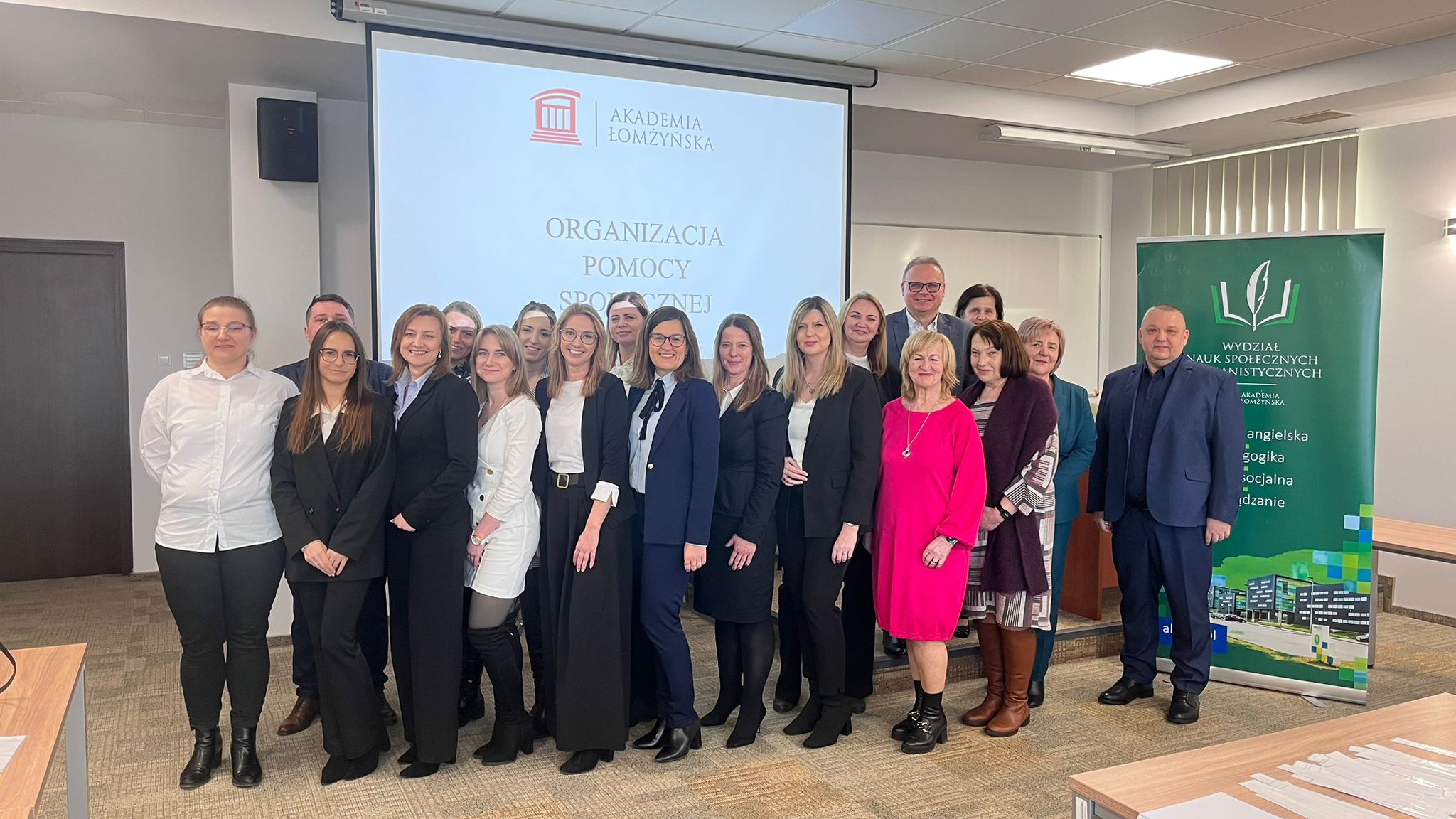 Duża grupa ludzi spozuje do zdjęcia. W tle na ekranie logo Akademii Łomżyńskiej i napis Organizacja pomocy społecznej. Widoczny rollup z napisem Wydział Nauk Społecznych i Humanistycznych
