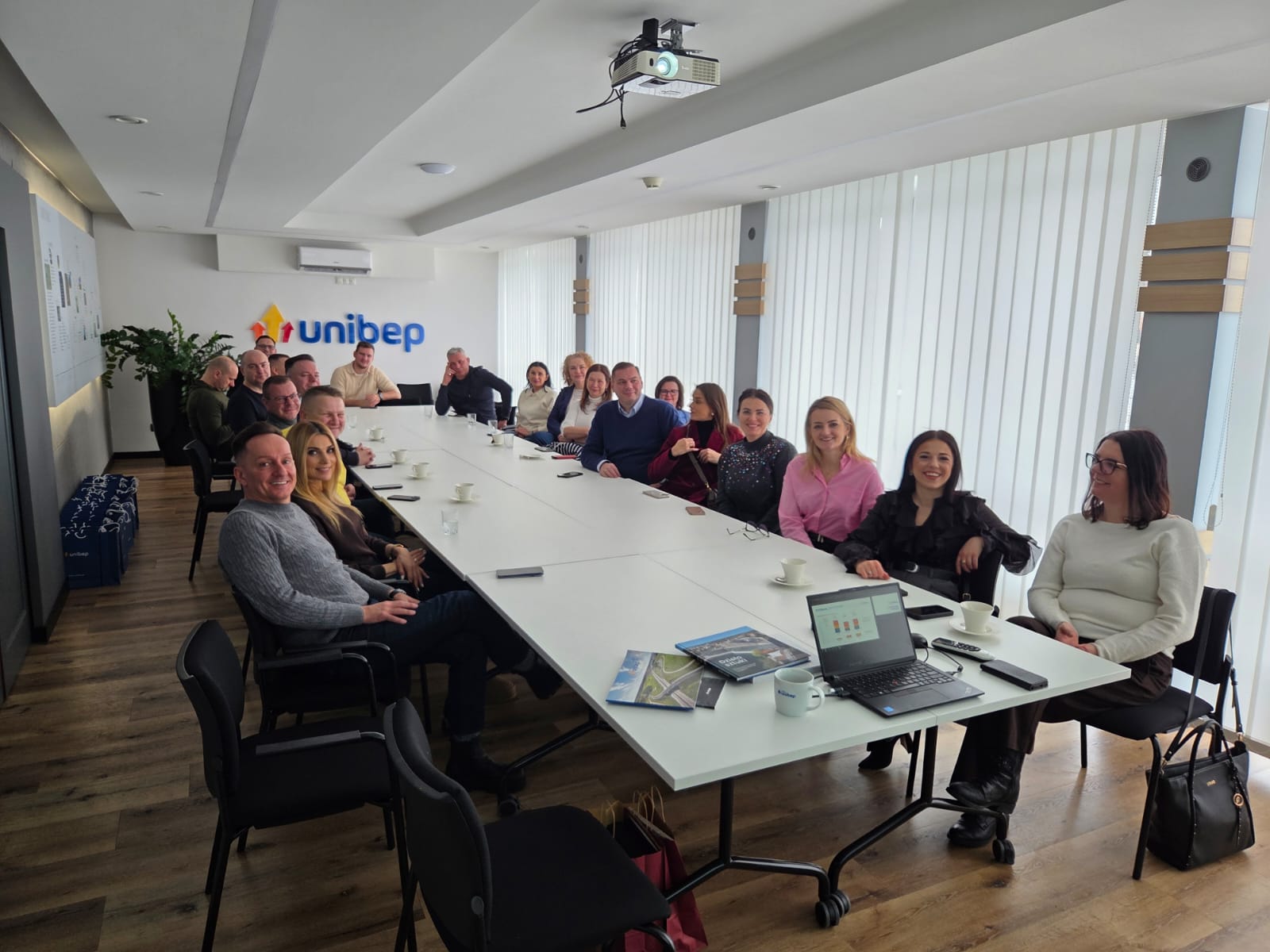 Duża grupa ludzi siedzi wokół podłużnego stołu. W drugim tle na ścianie logo unibep