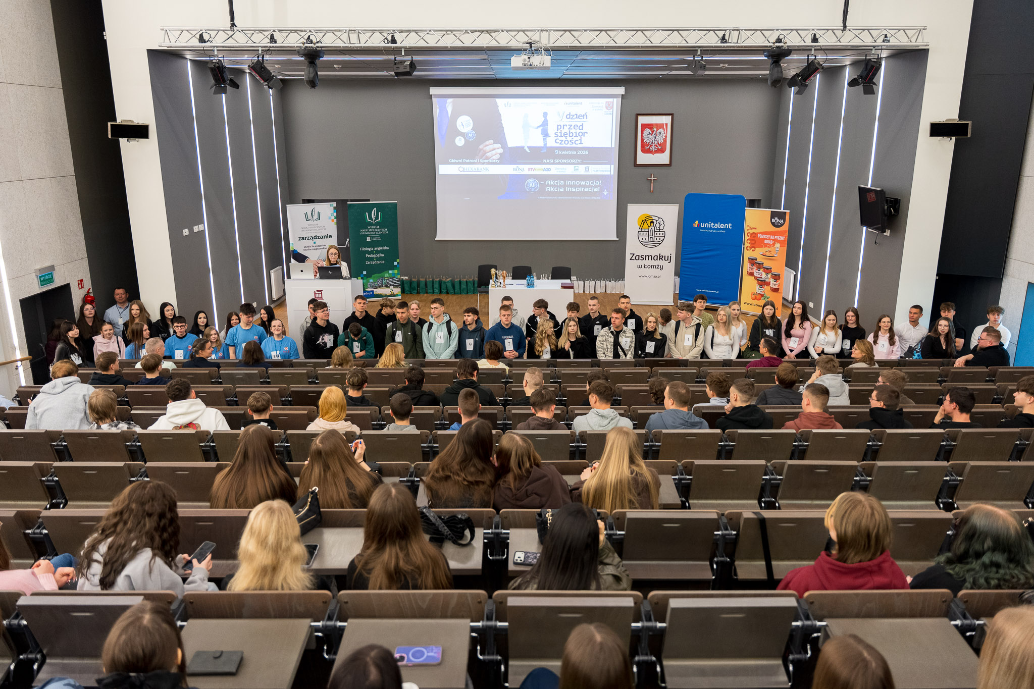 Zdjęcie przedstawia dużą aulę wykładową wypełnioną publicznością. Na pierwszym planie widoczne są rzędy siedzeń z osobami siedzącymi tyłem do aparatu. W środkowej i tylnej części sali znajdują się kolejne rzędy, w większości zajęte. Na scenie, przed zgromadzoną publicznością, stoi grupa kilkudziesięciu osób ustawionych w jednym rzędzie. Za nimi znajduje się długi stół konferencyjny oraz ekran projekcyjny z wyświetloną prezentacją. Po obu stronach sceny widoczne są pionowe banery z logotypami i grafikami. Nad sceną zamontowane jest oświetlenie sceniczne. Wnętrze sali jest nowoczesne, o jasnych ścianach i uporządkowanym układzie przestrzeni.