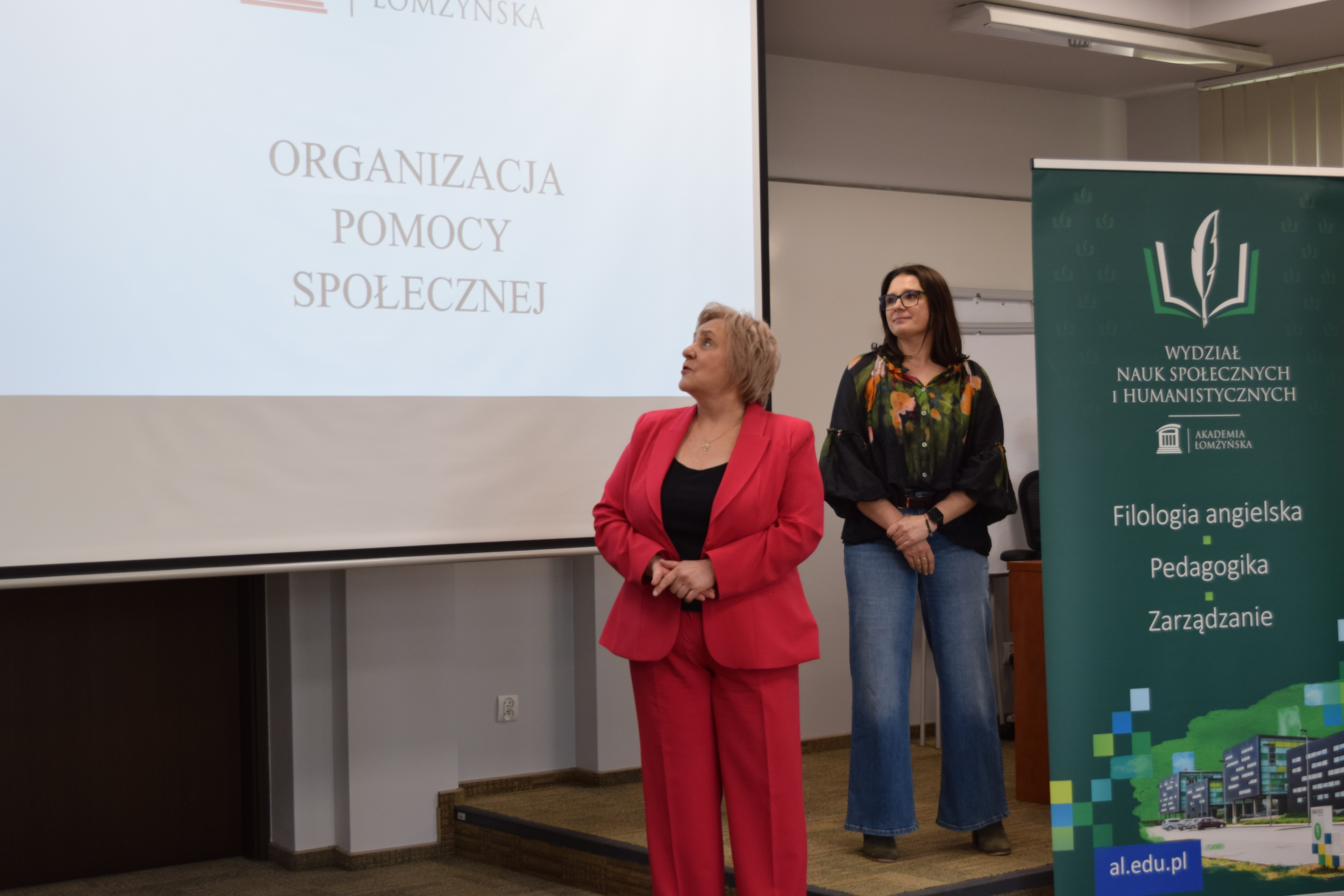 Dwie osoby stoją w sali wykładowej obok ekranu z prezentacją zawierającą tekst ‘Organizacja Pomocy Społecznej’. Po prawej stronie znajduje się roll‑up z nazwą Wydziału Nauk Społecznych i Humanistycznych oraz listą kierunków. Za osobami widoczna jest tablica i elementy sali.