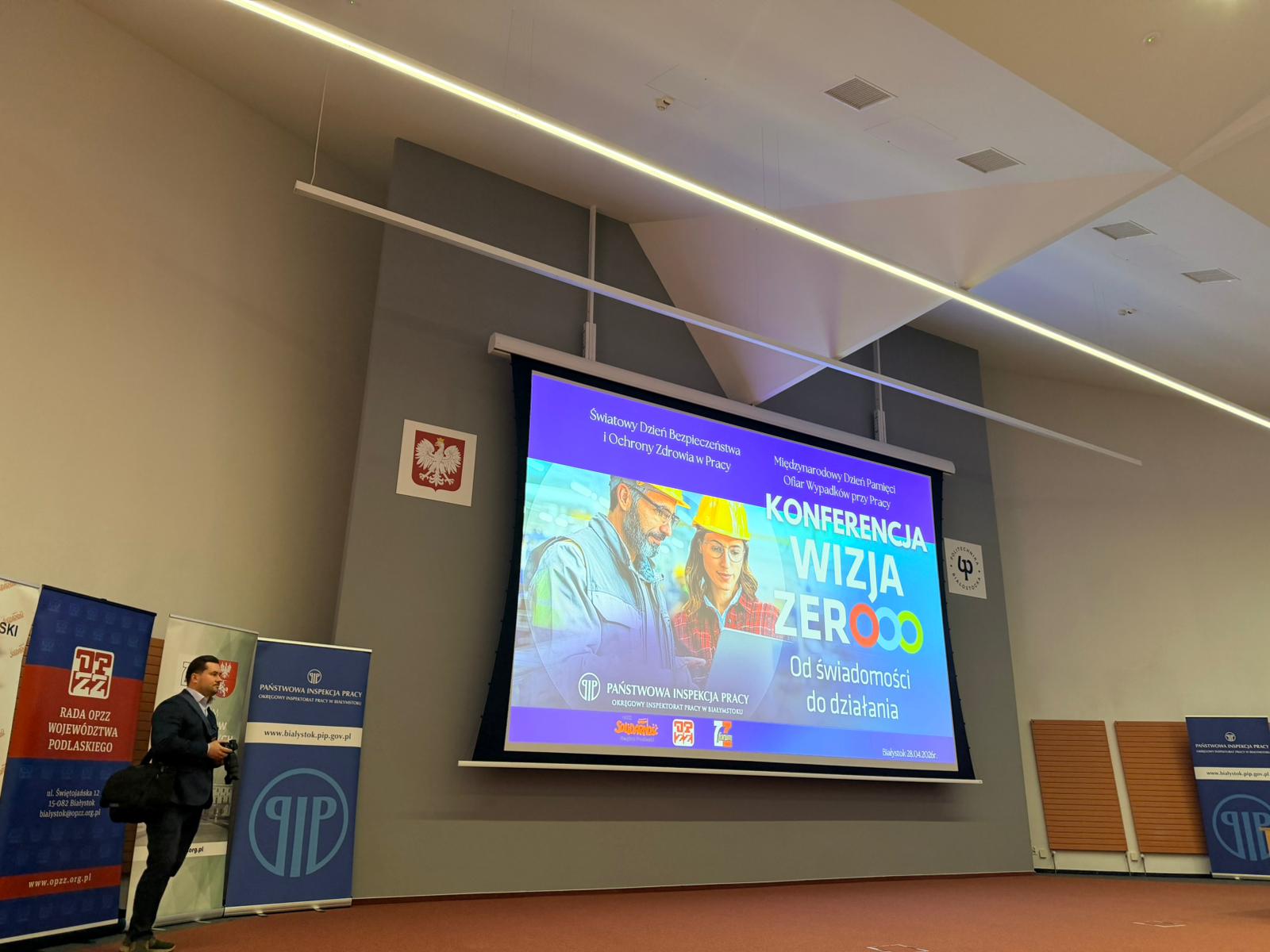 W sali konferencyjnej wyświetlony jest slajd konferencji Wizja Zero z grafiką osób w kaskach i informacjami o wydarzeniu, a obok stoją banery organizatorów oraz fotograf przygotowujący się do dokumentowania wydarzenia.