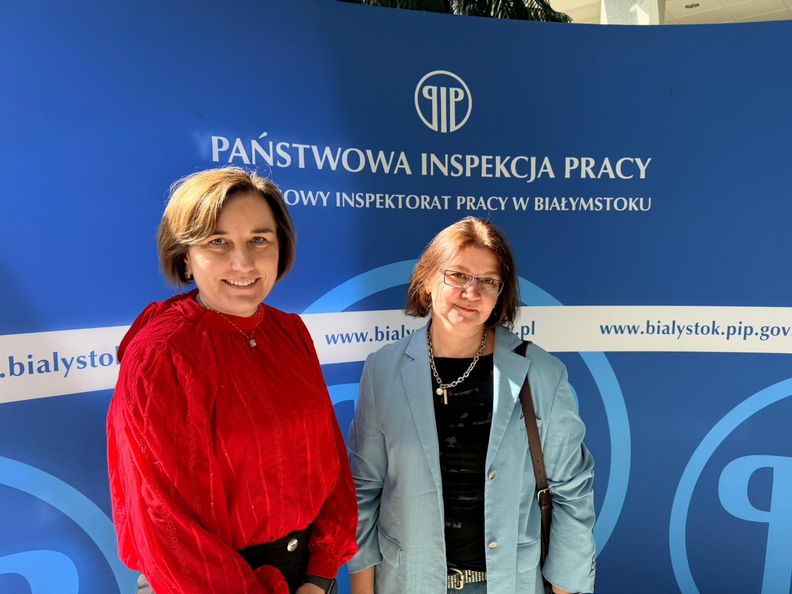 Panie Dziekan pozują do zdjęcia na tle niebieskiej ścianki z logo Państwowej Inspekcji Pracy.