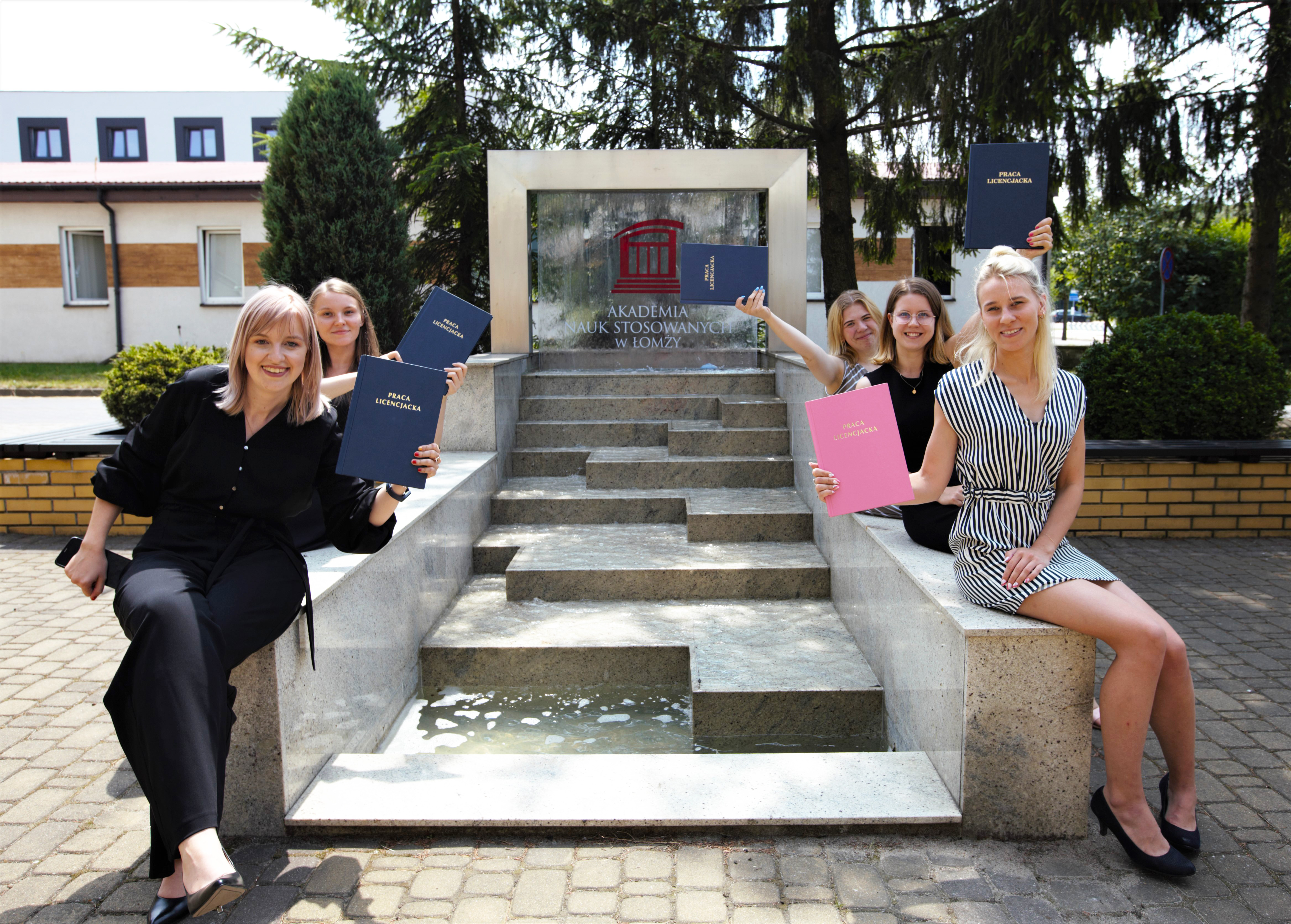 Studenci trzymają prace dyplomowe obok fontanny z logo Uczelni