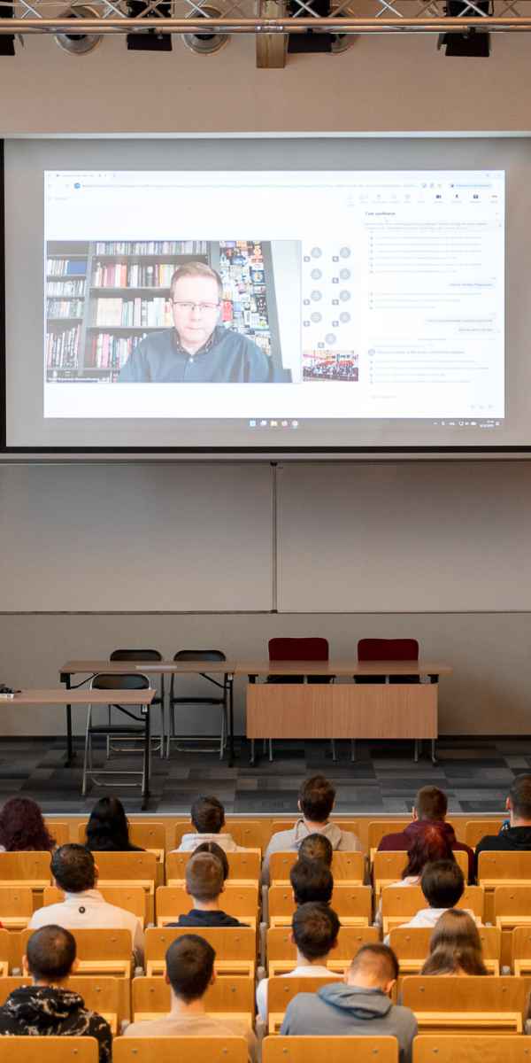 Ujęcie na scenę auli. Na pierwszym planie studenci siedzący na widowni. Na scenie po lewej stronie przy mównicy stoi dr Sitkowski. W centrum sceny na ekranie widoczny zaproszony gość