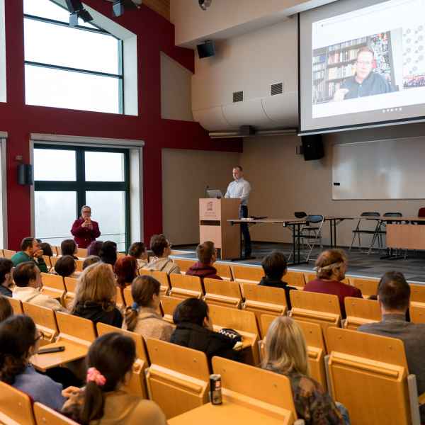 Grupa studentów siedzi w auli. Patrzą w kierunku sceny. Widoczna scena na której wyświetlona jest sylwetka gościa
