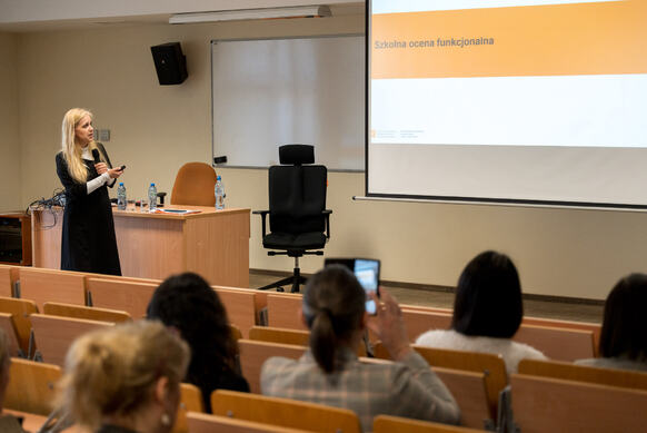Kobieta prowadzi prezentację w sali wykładowej, stojąc obok biurka, a na ekranie wyświetlony jest slajd zatytułowany ‘Szkolna ocena funkcjonalna’, podczas gdy uczestnicy siedzą w ławkach naprzeciwko.