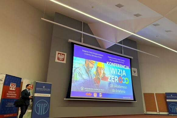 W sali konferencyjnej wyświetlony jest slajd konferencji Wizja Zero z grafiką osób w kaskach i informacjami o wydarzeniu, a obok stoją banery organizatorów oraz fotograf przygotowujący się do dokumentowania wydarzenia.