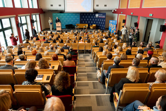 Duża sala wykładowa z licznie zgromadzonymi uczestnikami siedzącymi w rzędach, słuchającymi wystąpienia osoby stojącej przy mównicy na scenie