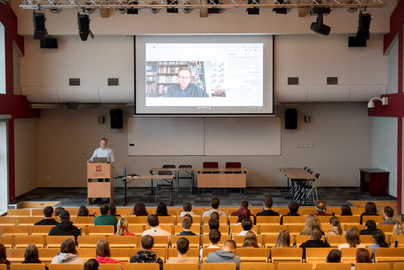 Ujęcie na scenę auli. Na pierwszym planie studenci siedzący na widowni. Na scenie po lewej stronie przy mównicy stoi dr Sitkowski. W centrum sceny na ekranie widoczny zaproszony gość