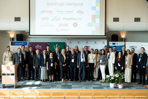Duża grupa uczestników konferencji pozuje na scenie. W tle slajd z widocznymi logówkami partnerów strategicznych