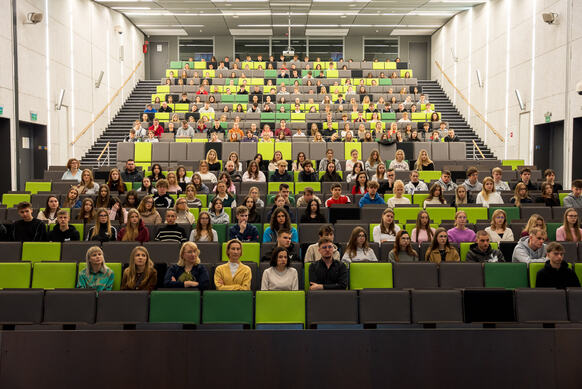 Duża aula wykładowa z wieloma rzędami zielono-szarych siedzeń, wypełniona uczestnikami wydarzenia.