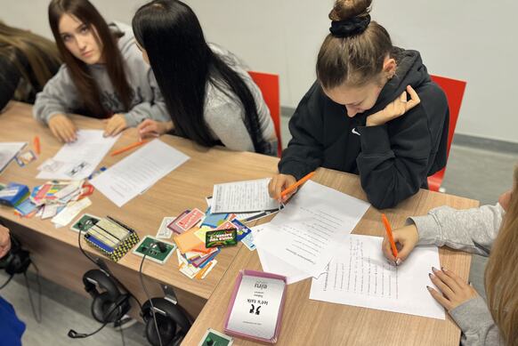 cztery młode dziewczyny siedzą przy ławkach i rozwiązują test językowy. Obok na blacie leżą kolorowe karty do gry z napisami po angielsku