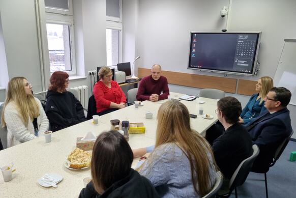 Przy długim stole siedzą studenci i pracownicy instytucji. Jedna z osób mówi