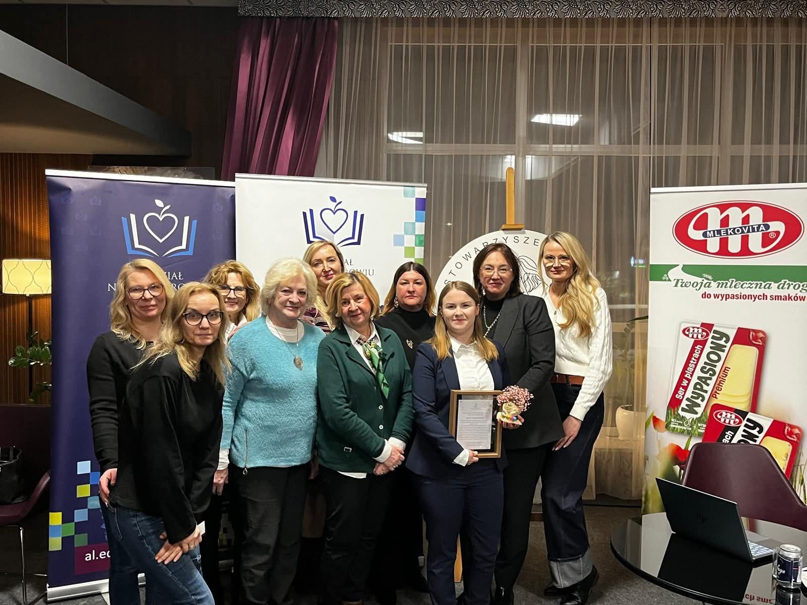 Grupa uczestniczek wydarzenia pozuje wspólnie do zdjęcia na tle roll upów Wydziału Nauk o Zdrowiu, kierunku kosmetologia oraz baneru sponsorskiego. Jedna z kobiet trzyma wręczone podziękowanie i bukiet kwiatów