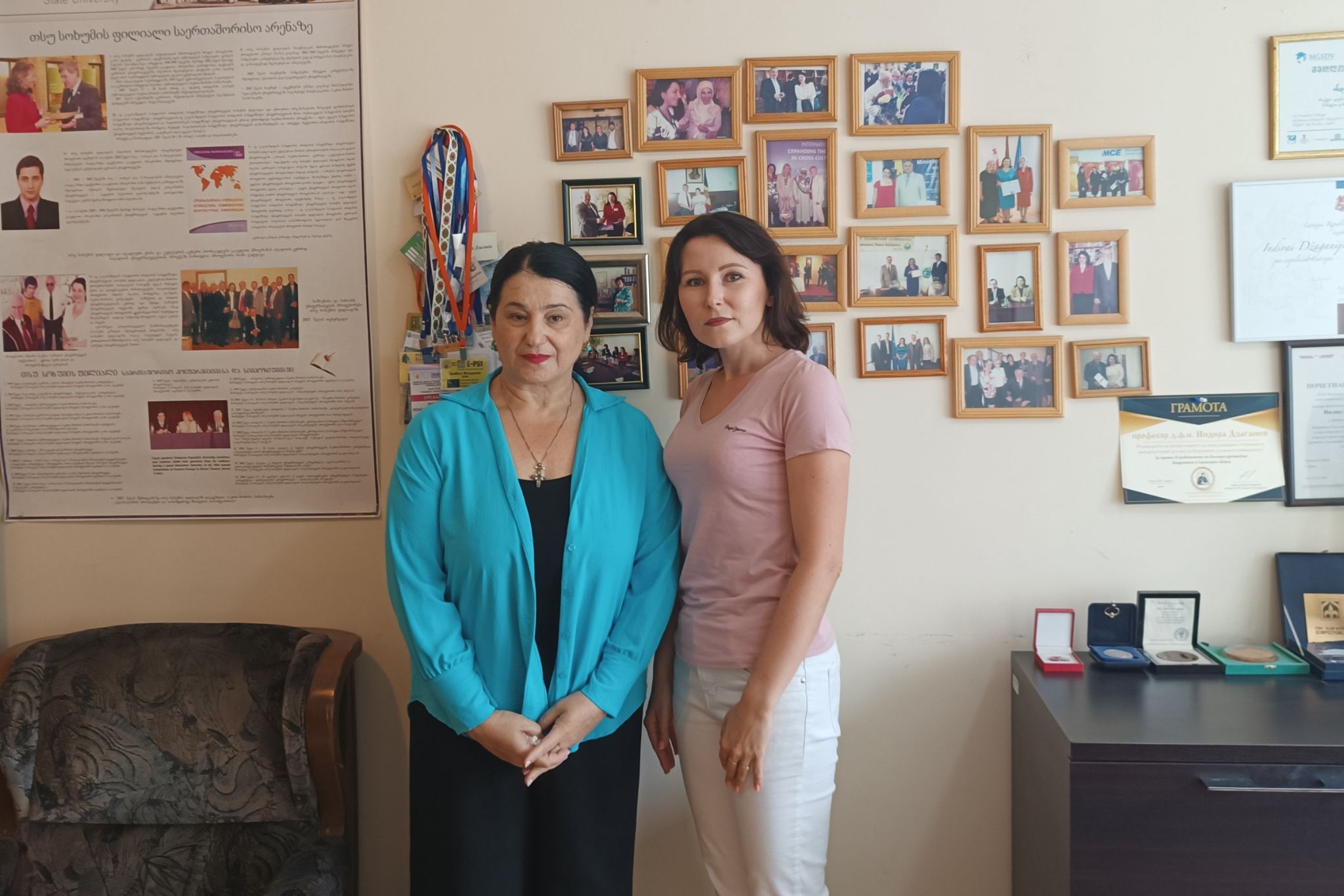 Dwie osoby stoją obok siebie w biurze lub gabinecie. W tle na jasnej ścianie znajduje się galeria licznych oprawionych fotografii oraz kilka dyplomów i certyfikatów. Po lewej stronie widoczny jest duży plakat informacyjny oraz fotel, a po prawej komoda z medalami i pamiątkowymi odznaczeniami w etui. Jedna osoba ma turkusowy kardigan i czarną suknię, druga jasnoróżową koszulkę i białe spodnie. Zdjęcie wykonano w dobrze oświetlonym wnętrzu o charakterze akademickim lub administracyjnym.