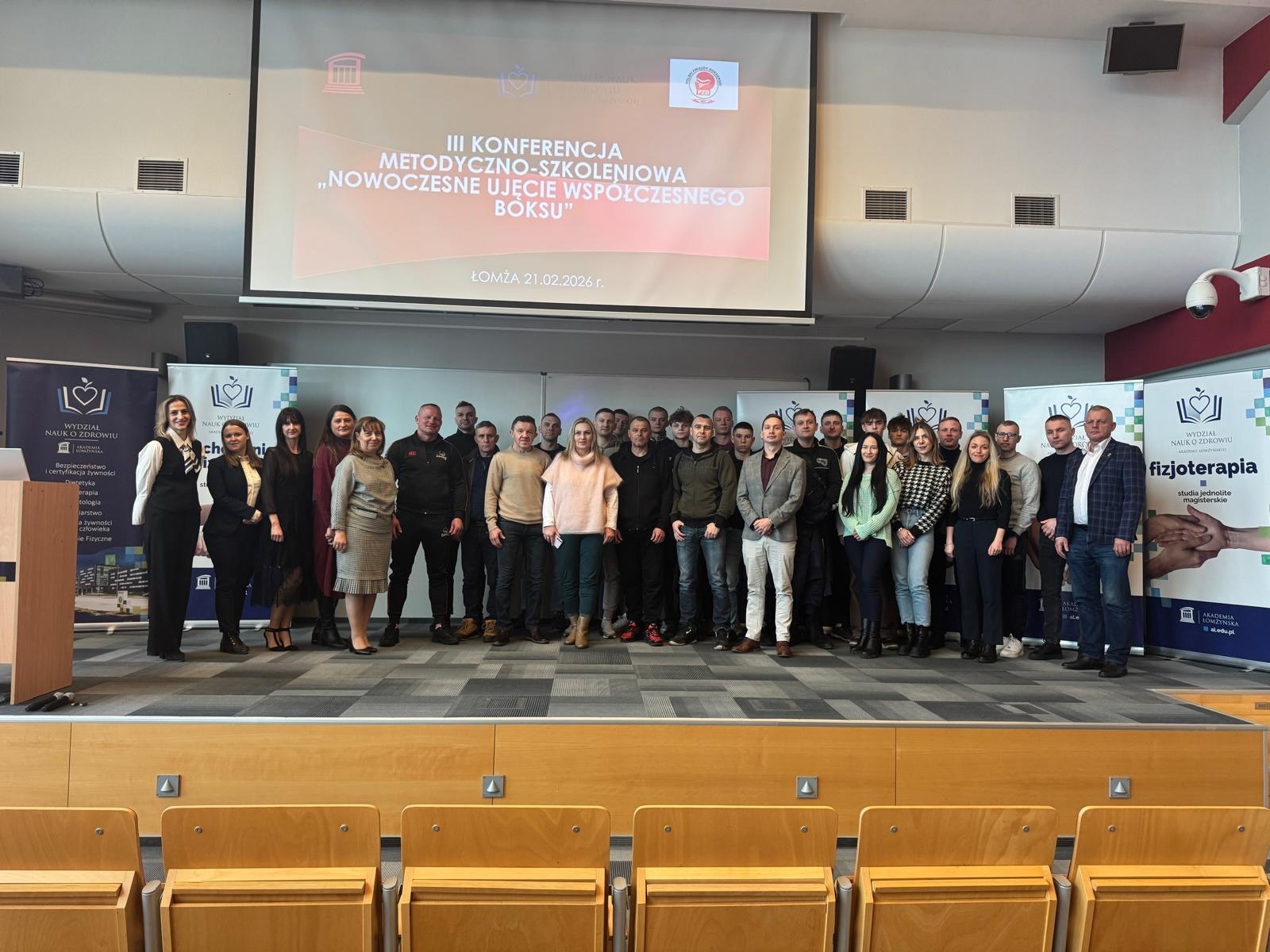 Duża grupa uczestników stoi na scenie przed ekranem projekcyjnym. Na ekranie widoczny jest tytuł konferencji „III Konferencja Metodyczno‑Szkoleniowa – Nowoczesne Ujęcie Współczesnego Boksu”. Po bokach znajdują się roll‑upy z logo uczelni. Przed grupą widać puste rzędy krzeseł.