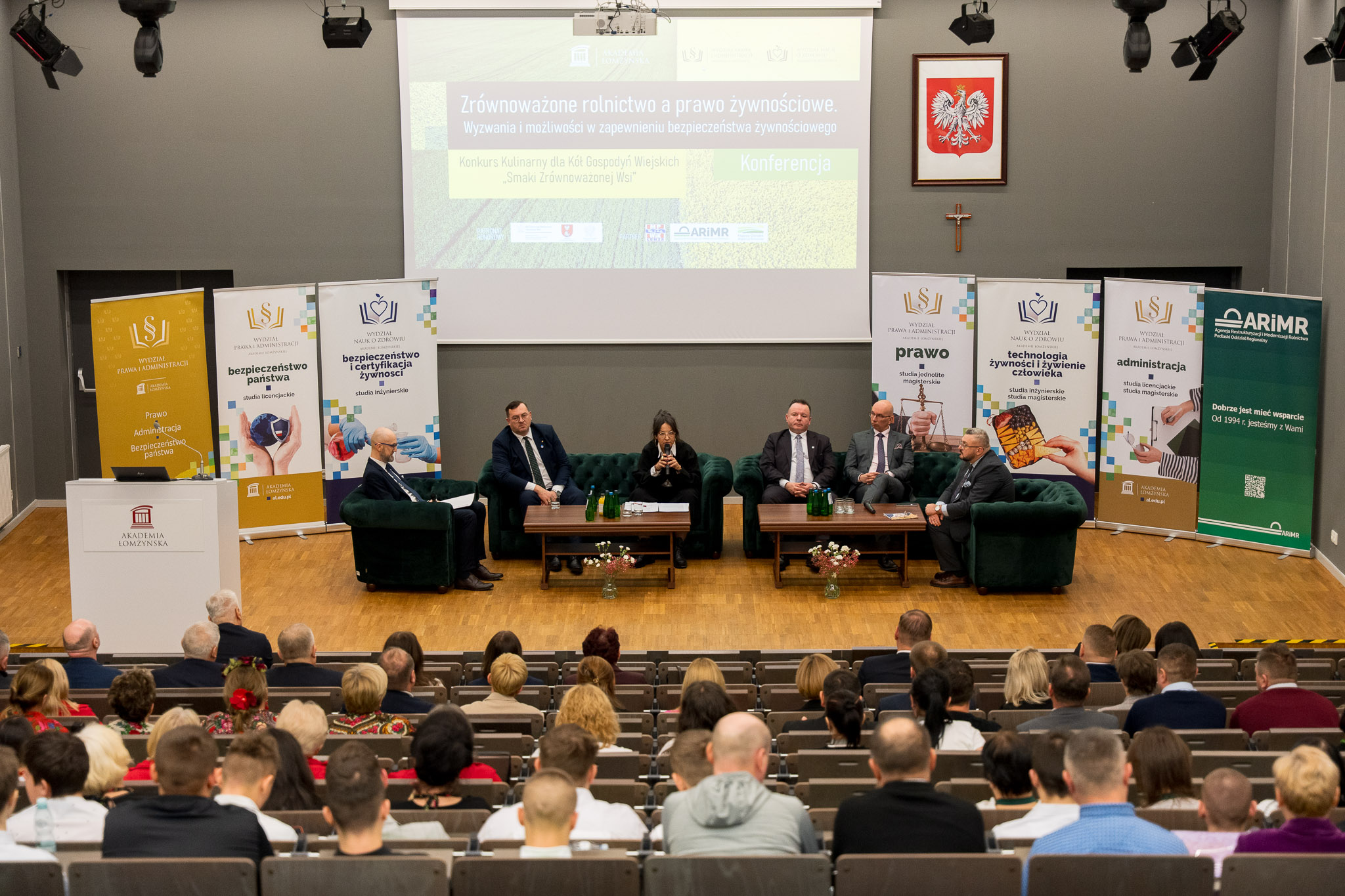 Widok z tyłu sali konferencyjnej, w której uczestnicy siedzą w kilku rzędach foteli ustawionych przed sceną. Na scenie znajduje się sześć osób siedzących w zielonych fotelach wokół niskich stolików. Za nimi widoczny jest duży ekran z prezentacją zatytułowaną ‘Zrównoważone rolnictwo a prawo żywnościowe’. Po obu stronach sceny ustawiono liczne roll upy przedstawiające logotypy oraz treści związane z tematyką wydarzenia. Nad ekranem wisi godło państwowe. W sali panuje jasne oświetlenie, a uczestnicy uważnie obserwują prelegentów