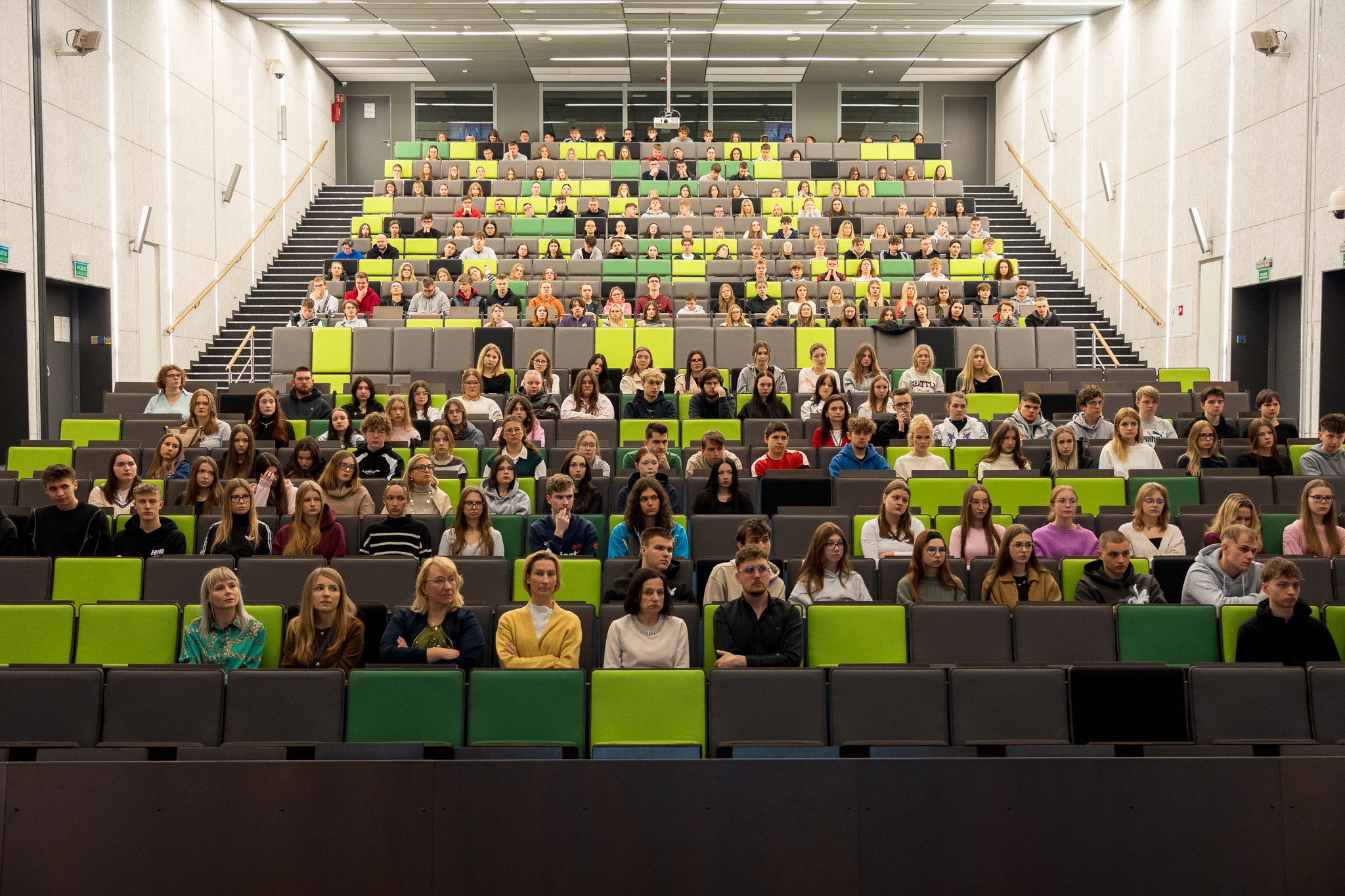 Duża grupa uczniów i uczennic siedzi w nowoczesnej, przestronnej auli wykładowej. Miejsca są ułożone amfiteatralnie, w naprzemiennych rzędach zielonych i szarych foteli. Sala jest dobrze oświetlona, a na ścianach znajdują się minimalistyczne panele akustyczne. Uczniowie zajmują niemal wszystkie miejsca i patrzą w stronę aparatu, tworząc wrażenie pełnej, intensywnie uczestniczącej publiczności.