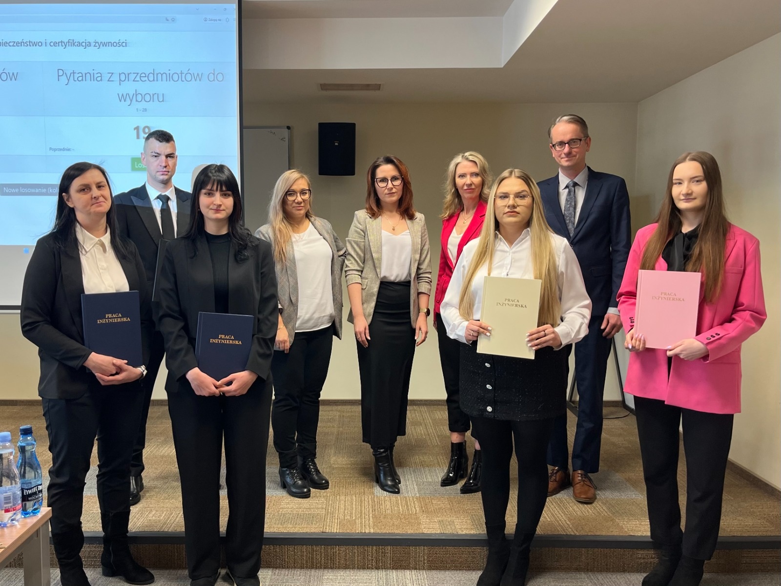 Grupa osób stoi na tle sali wykładowej przed ekranem projekcyjnym. Kilka osób w pierwszym rzędzie trzyma teczki lub dyplomy w różnych kolorach. Wszyscy są ubrani w eleganckie stroje, a ustawienie sugeruje zakończenie wydarzenia, takiego jak obrona prac lub wręczenie dokumentów. Na stole z boku widoczne są butelki wody, a w tle znajduje się prezentacja wyświetlana na ekranie.