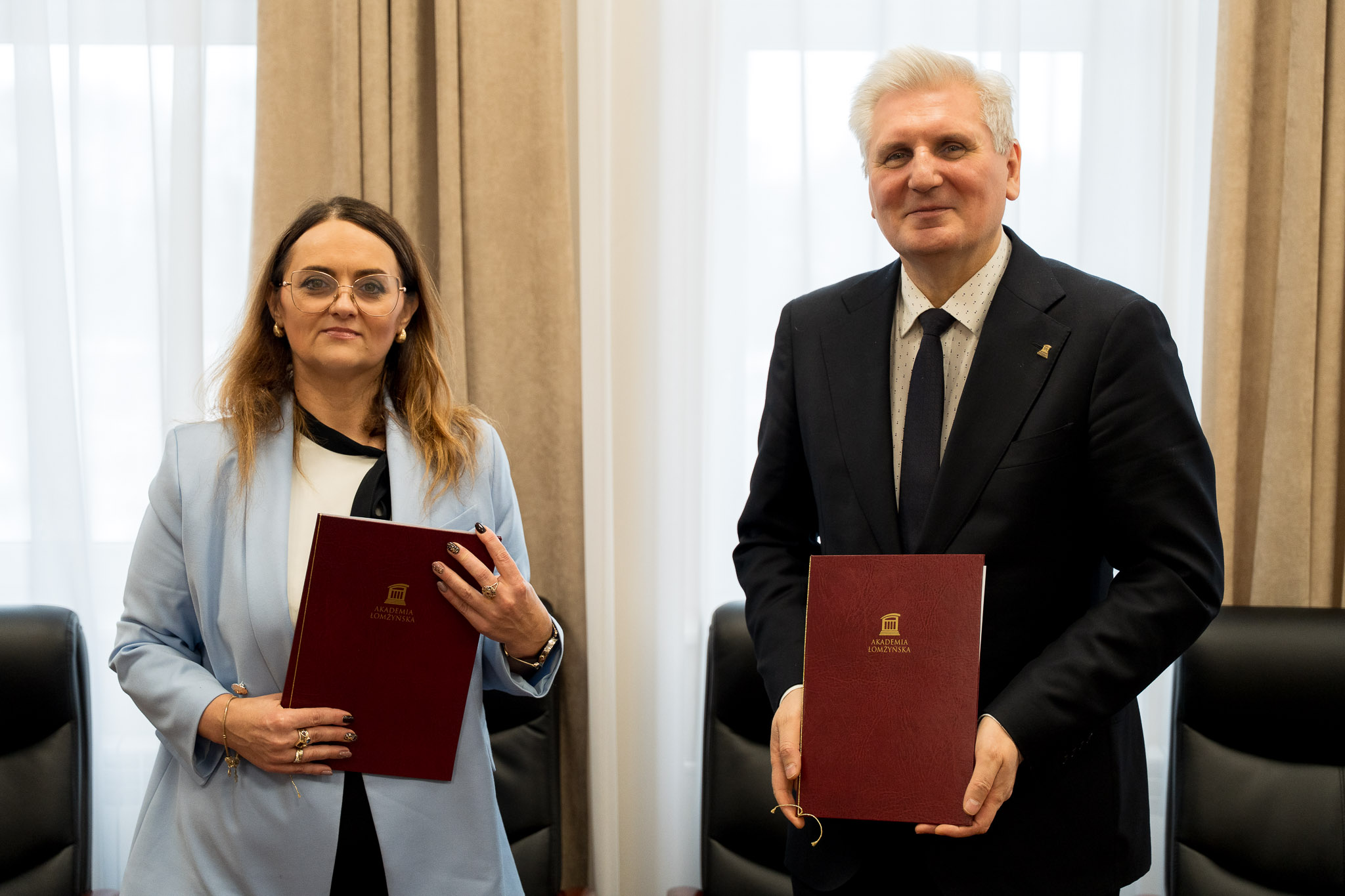Dwie osoby stoją obok siebie w eleganckim, jasnym pomieszczeniu. Każda z nich trzyma w rękach bordową teczkę z logo Akademii Łomżyńskiej. Jedna osoba ma na sobie jasnoniebieską marynarkę, a druga ciemny garnitur z krawatem. W tle widoczne są zasłony oraz fragment skórzanych krzeseł.