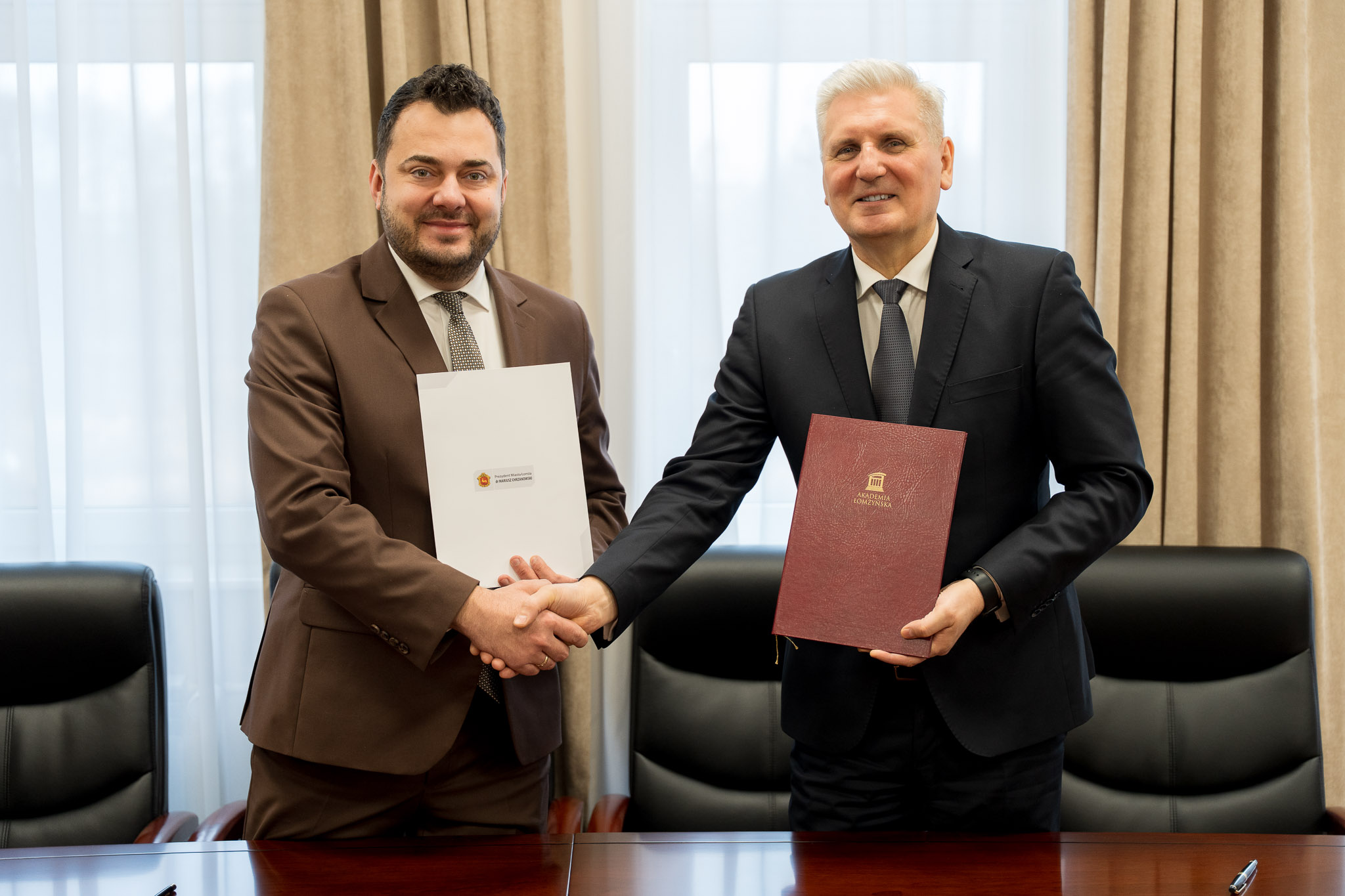 Dwie osoby w formalnych strojach stoją przy stole konferencyjnym, podając sobie ręce w geście zawarcia porozumienia. Jedna z osób trzyma teczkę w bordowym kolorze z logo uczelni, a druga trzyma biały dokument z widocznym logo instytucji. W tle znajdują się beżowe zasłony oraz wysokie okno.