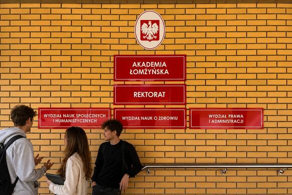 Trzy osoby stoją przy ścianie z jasnej cegły wewnątrz budynku uczelni. Na ścianie widoczne są czerwone tablice informacyjne z napisami: „Akademia Łomżyńska”, „Rektorat”, „Wydział Nauk Społecznych i Humanistycznych”, „Wydział Nauk o Zdrowiu” oraz „Wydział Prawa i Administracji”. Nad tablicami znajduje się godło Polski. W dolnej części ściany zamontowana jest metalowa poręcz.