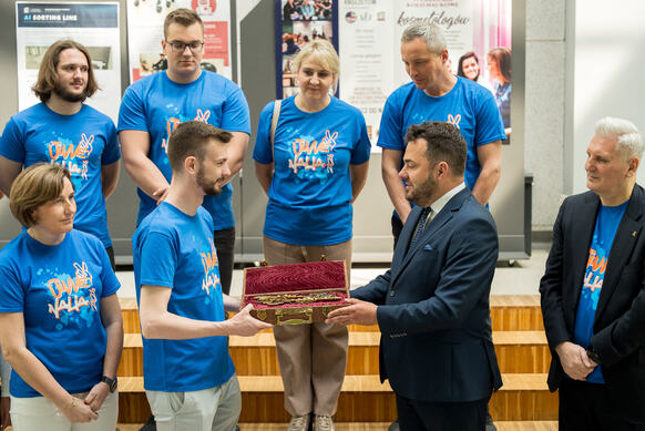 Grupa osób stojących w pomieszczeniu przypominającym salę uczelni lub instytucji. Kilka osób w niebieskich koszulkach z nadrukiem „Dni Nauki” otacza osobę w stroju formalnym, która wręcza ozdobną szkatułkę z dużym, dekoracyjnym kluczem. W tle widoczne są plakaty informacyjne i prezentacyjne, a uczestnicy stoją na podwyższeniu i wokół niego, obserwując przekazanie przedmiotu.