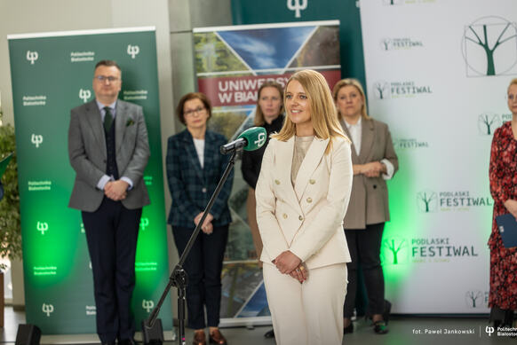 Osoba stojąca przy mikrofonie przemawia podczas wydarzenia; w tle widoczna jest grupa uczestników oraz banery z logotypami „Podlaski Festiwal Nauki i Sztuki”, „Uniwersytet w Białymstoku” i „Politechnika Białostocka”.