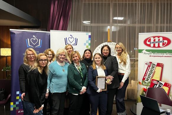 Grupa uczestniczek wydarzenia pozuje wspólnie do zdjęcia na tle roll upów Wydziału Nauk o Zdrowiu, kierunku kosmetologia oraz baneru sponsorskiego. Jedna z kobiet trzyma wręczone podziękowanie i bukiet kwiatów