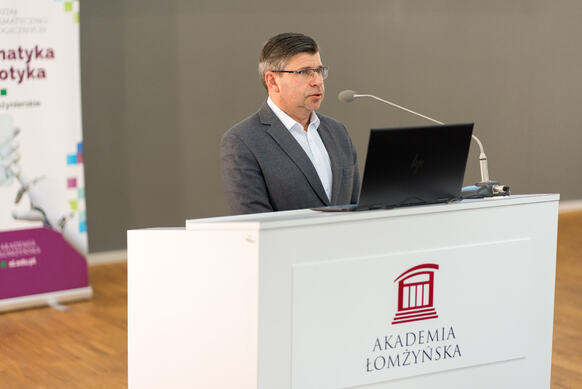 Osoba stoi za białą mównicą z logo Akademii Łomżyńskiej i przemawia do publiczności, korzystając z laptopa oraz mikrofonu. W tle widoczny jest roll up promujący informatykę oraz robotykę.