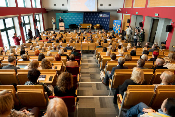 Widok z tyłu dużej sali audytoryjnej wypełnionej uczestnikami siedzącymi w rzędach drewnianych krzeseł. Na scenie z przodu stoi osoba przemawiająca przy mównicy, obok której znajdują się dwa fotele i stolik. W tle widoczna jest niebieska ścianka z logo instytucji, a z prawej strony ustawiony jest zespół kamer rejestrujących wydarzenie. Po lewej stronie sali widać duże okna sięgające od podłogi do sufitu, wpuszczające naturalne światło.