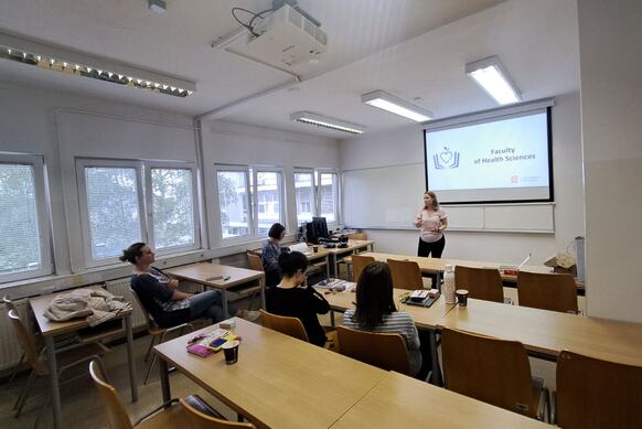 Sala wykładowa z rzędami drewnianych ławek i dużymi oknami po lewej stronie. Z przodu stoi osoba prowadząca prezentację, obok opuszczony ekran z napisem „Faculty of Health Sciences” oraz logo uczelni. Kilka osób siedzi przy stołach i słucha, na blacie widoczne są zeszyty, kubki i przybory. Na suficie zamontowany jest projektor i oświetlenie jarzeniowe.