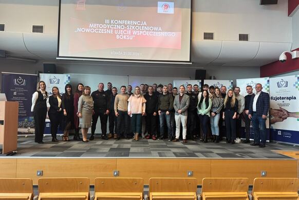 Duża grupa uczestników stoi na scenie przed ekranem projekcyjnym. Na ekranie widoczny jest tytuł konferencji „III Konferencja Metodyczno‑Szkoleniowa – Nowoczesne Ujęcie Współczesnego Boksu”. Po bokach znajdują się roll‑upy z logo uczelni. Przed grupą widać puste rzędy krzeseł.