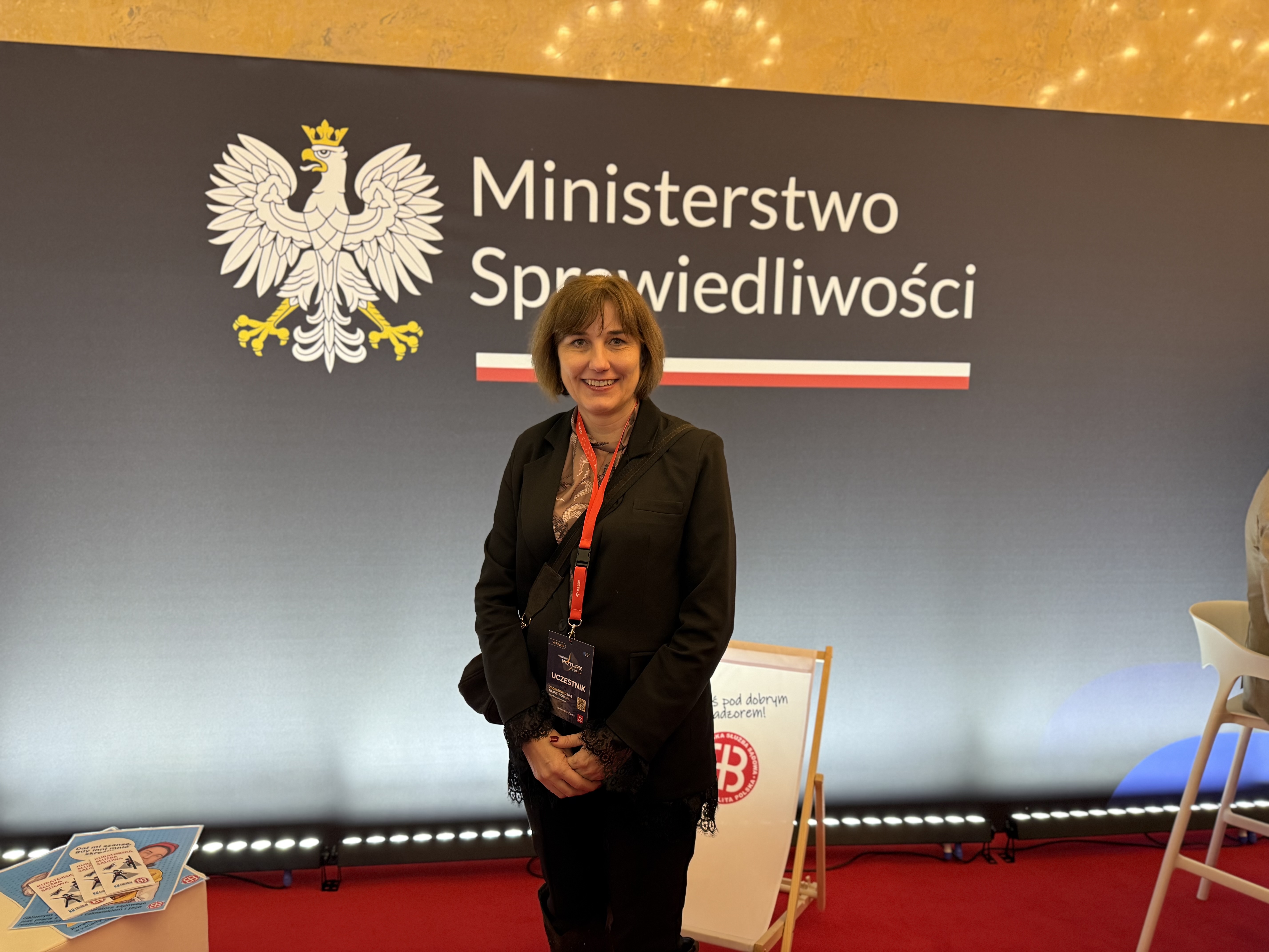 Kobieta z identyfikatorem pozuje na tle napisu Ministerstwo Sprawiedliwości z grafiką godła Państwa Polskiego.