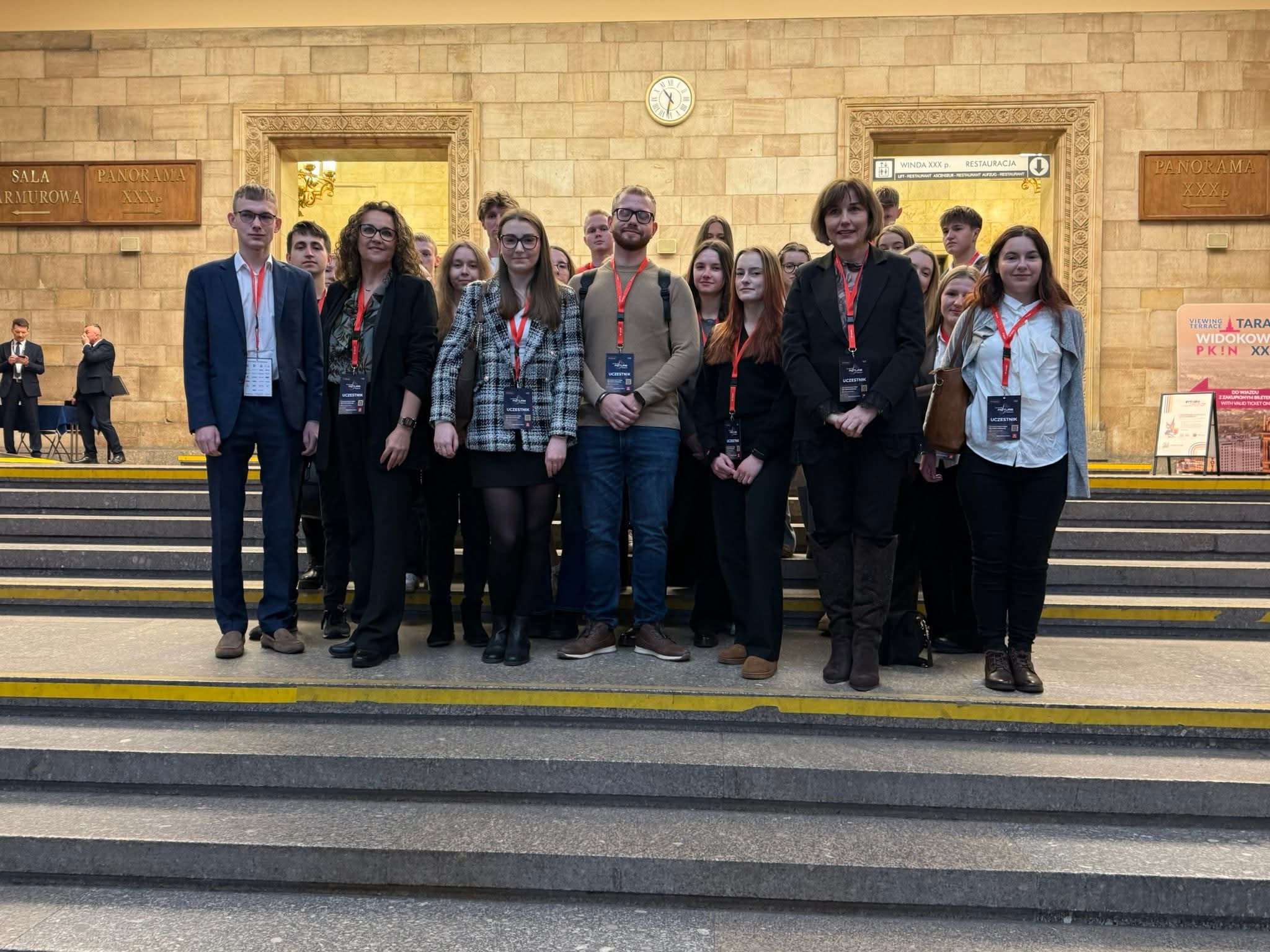 Grupa osób na schodach wewnątrz budynku o kamiennej aranżacji. Uczestnicy noszą identyfikatory, co sugeruje wydarzenie konferencyjne.