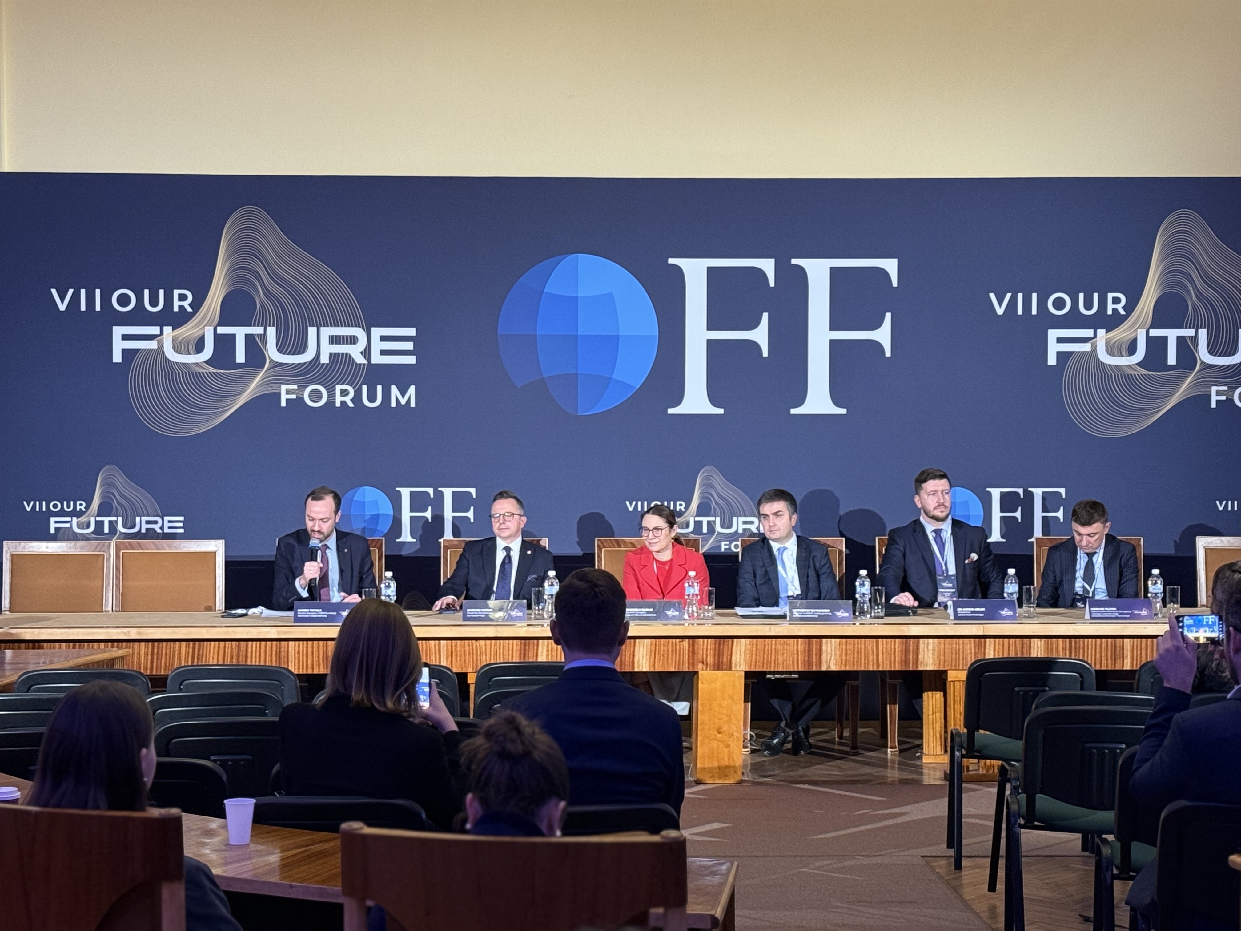 Grupa kilku osób siedzących za stołem konferencyjnym przemawia do osób siedzących na krzesłach.