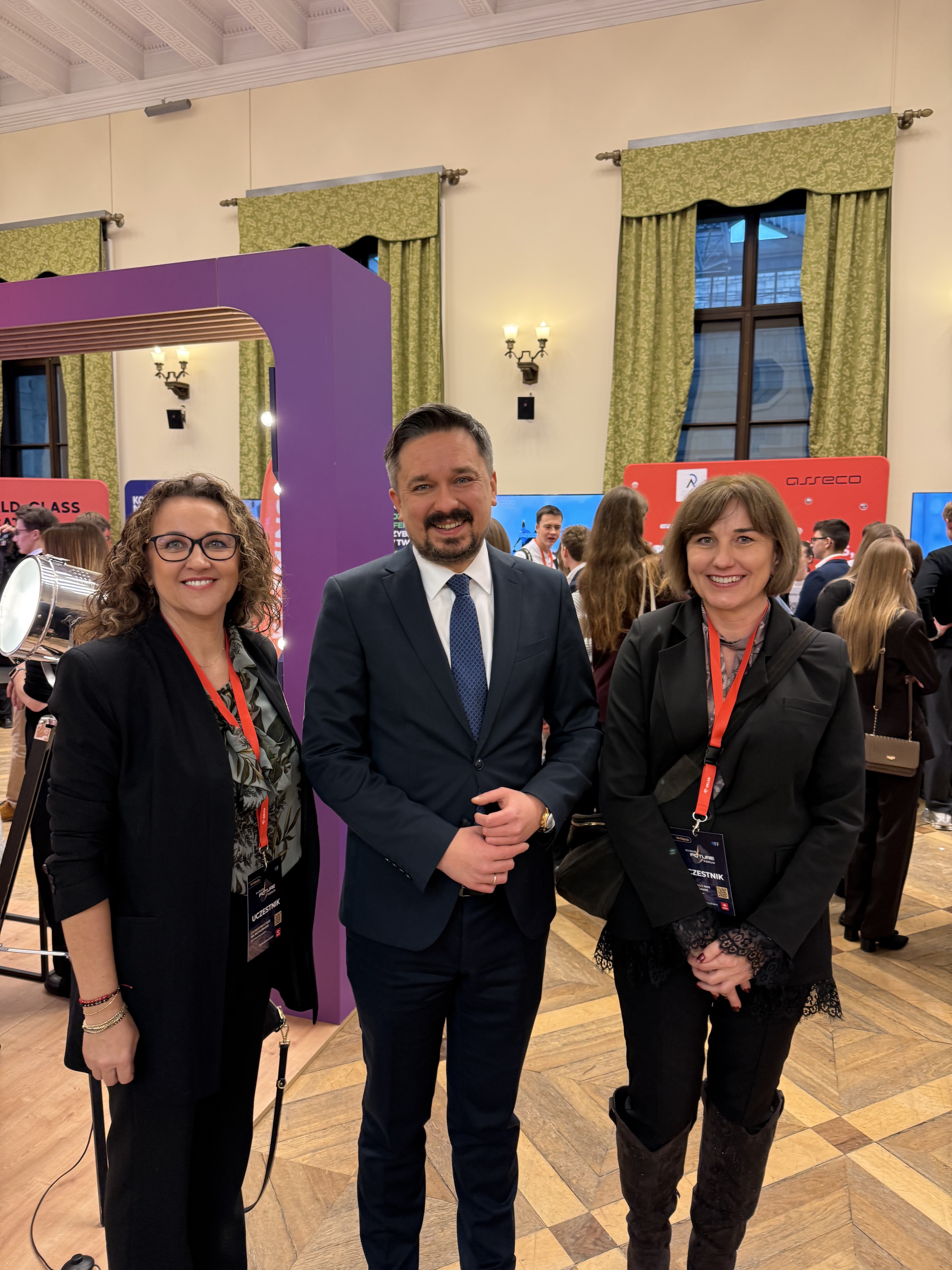 Dwie kobiety i mężczyzna pozują do zdjęcia na tle ściany z oknami i grupki innych osób.